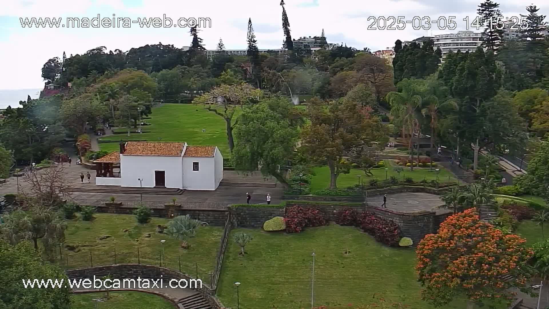 An aerial view of a park with a white building, lush green lawns, flowering trees, and a few people walking around on a partly cloudy day.