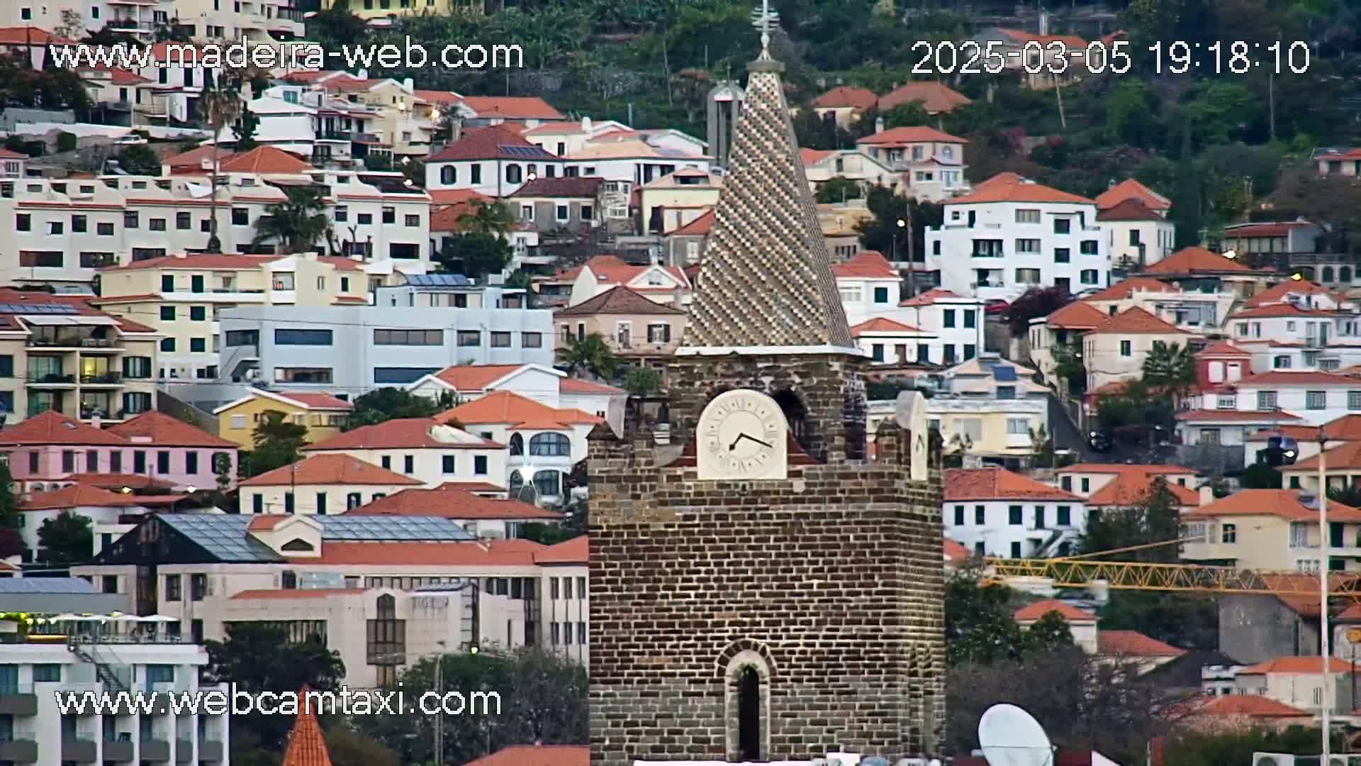 A brick clock tower stands prominently amidst a densely packed hillside of red-roofed buildings, under overcast skies.