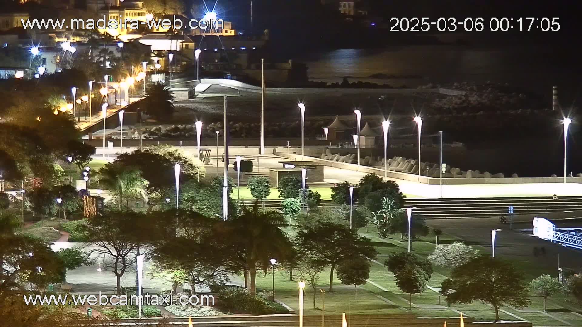 A nighttime view of a park-like area next to a body of water, illuminated by numerous tall lampposts.