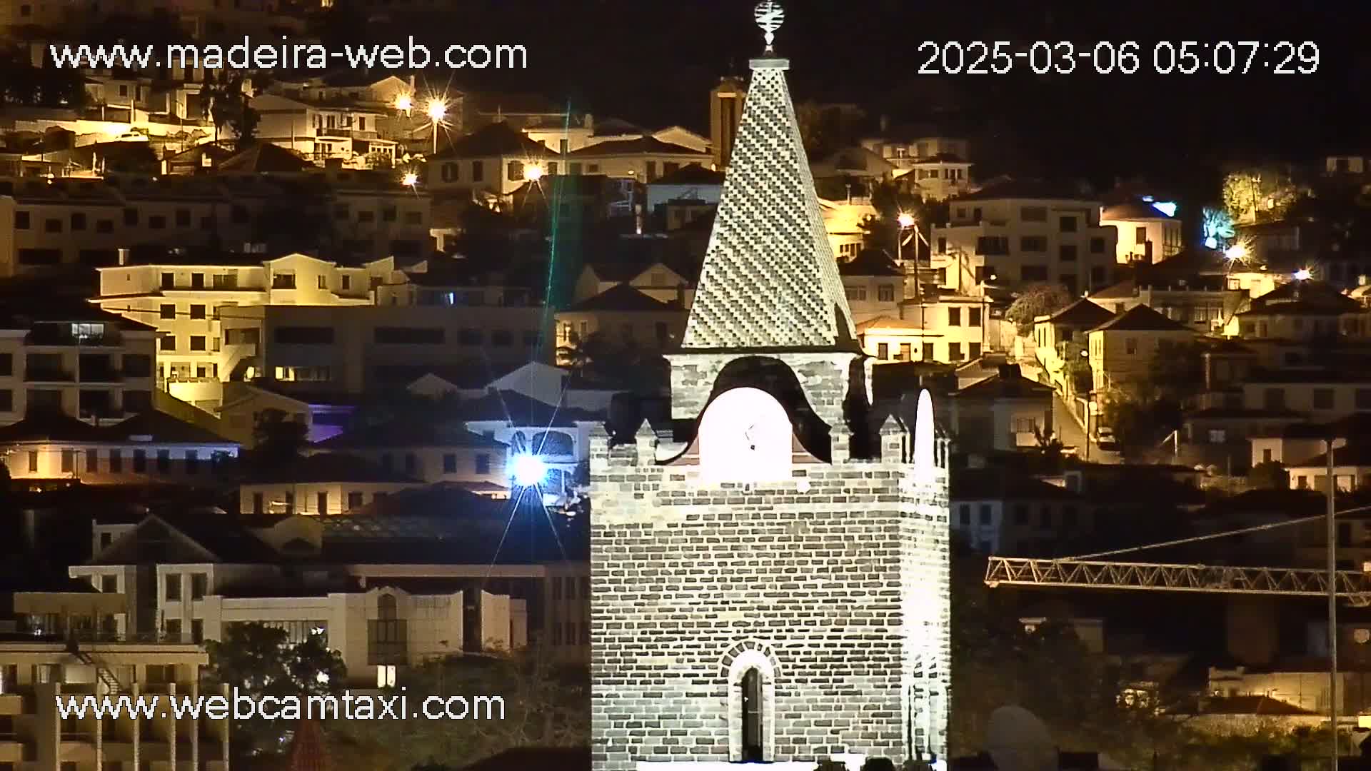 A stone church tower is illuminated at night, overlooking a hillside town with many brightly lit buildings.