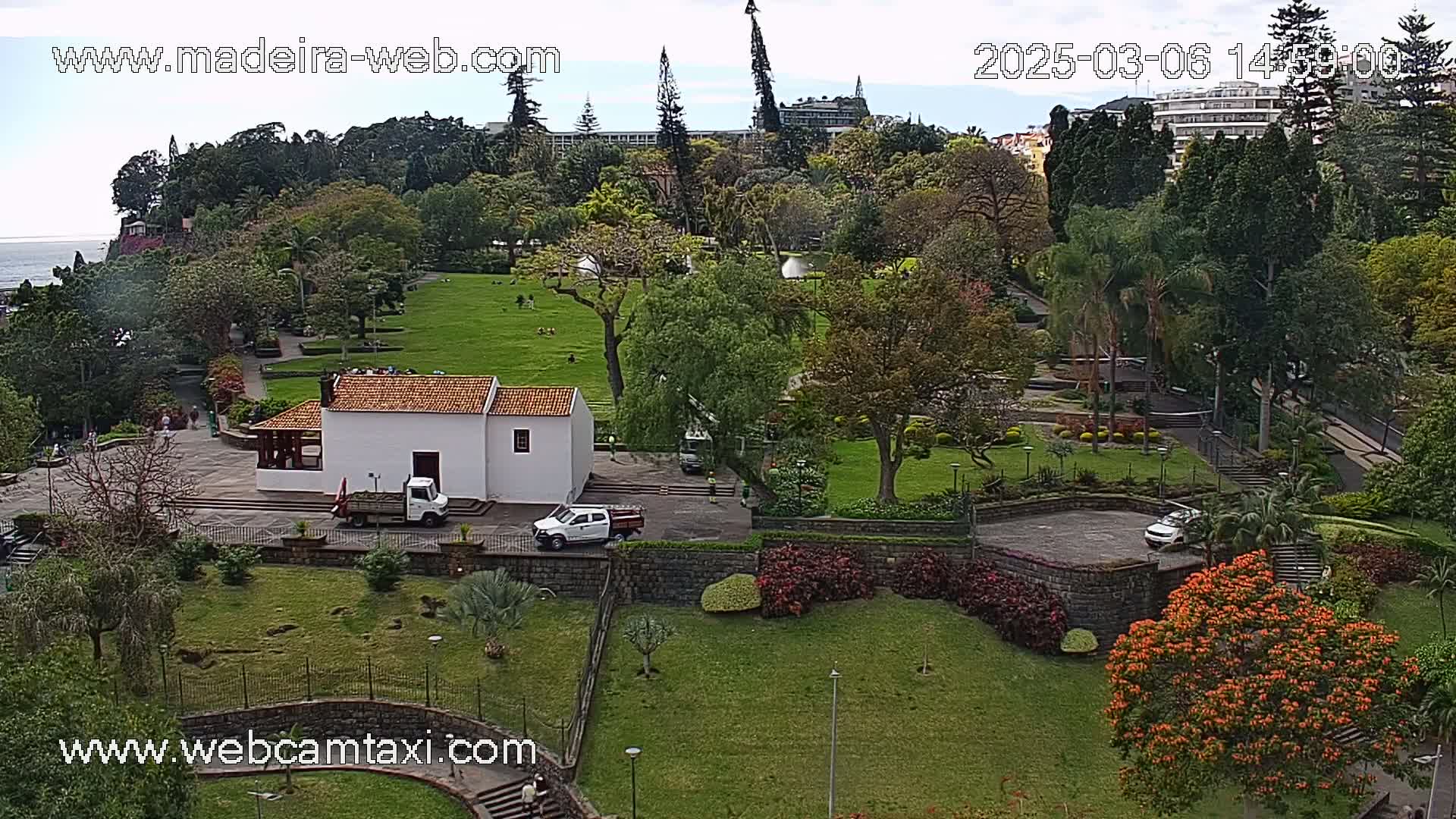 A mostly sunny aerial view of a park with a white building, several vehicles, and lush green lawns and trees, overlooking a coastline.
