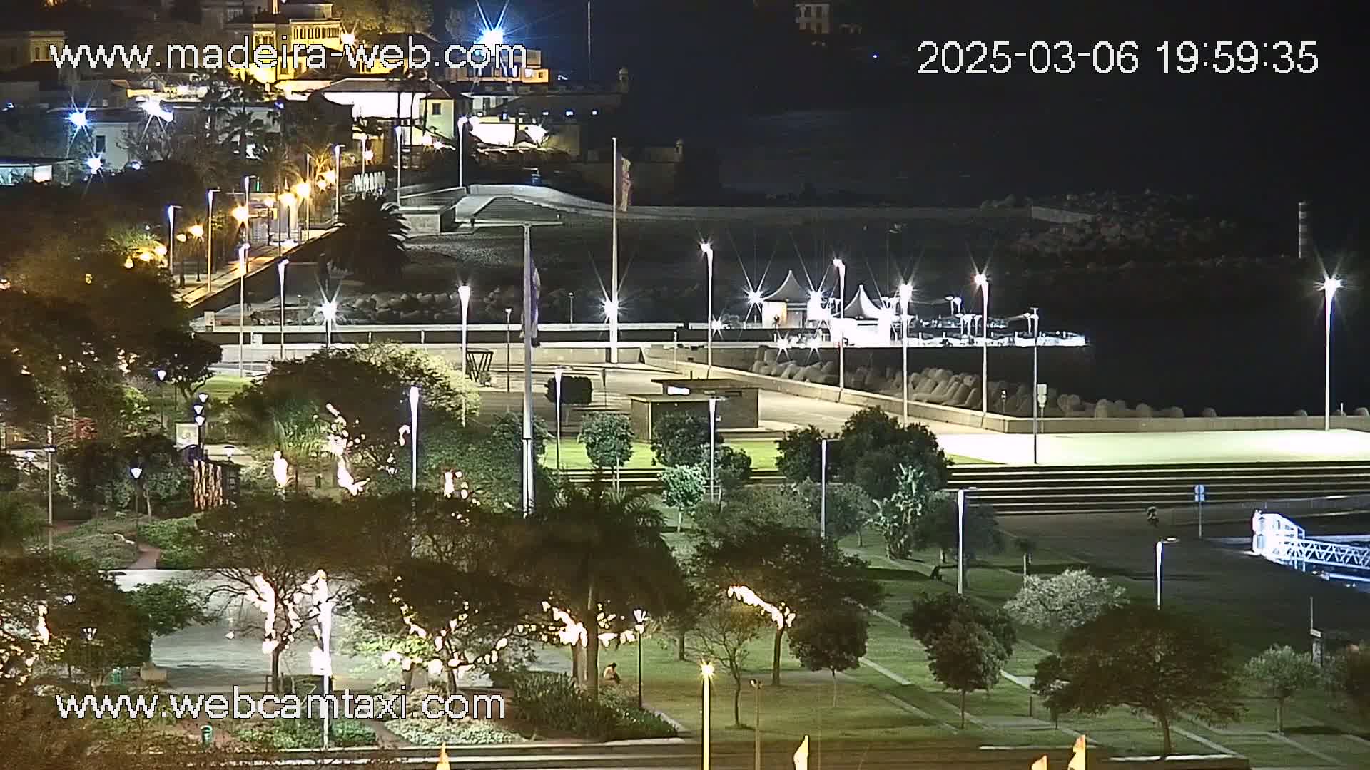 A nighttime view of a park and waterfront area, illuminated by numerous lights, with a structure resembling a pavilion near the water's edge.
