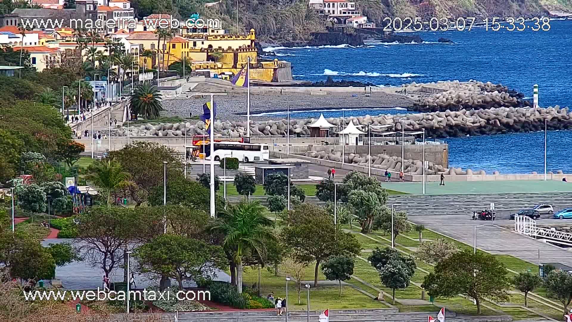 A coastal town scene with a park, a bus, and a pebble beach bordered by a breakwater, under a mostly sunny sky.
