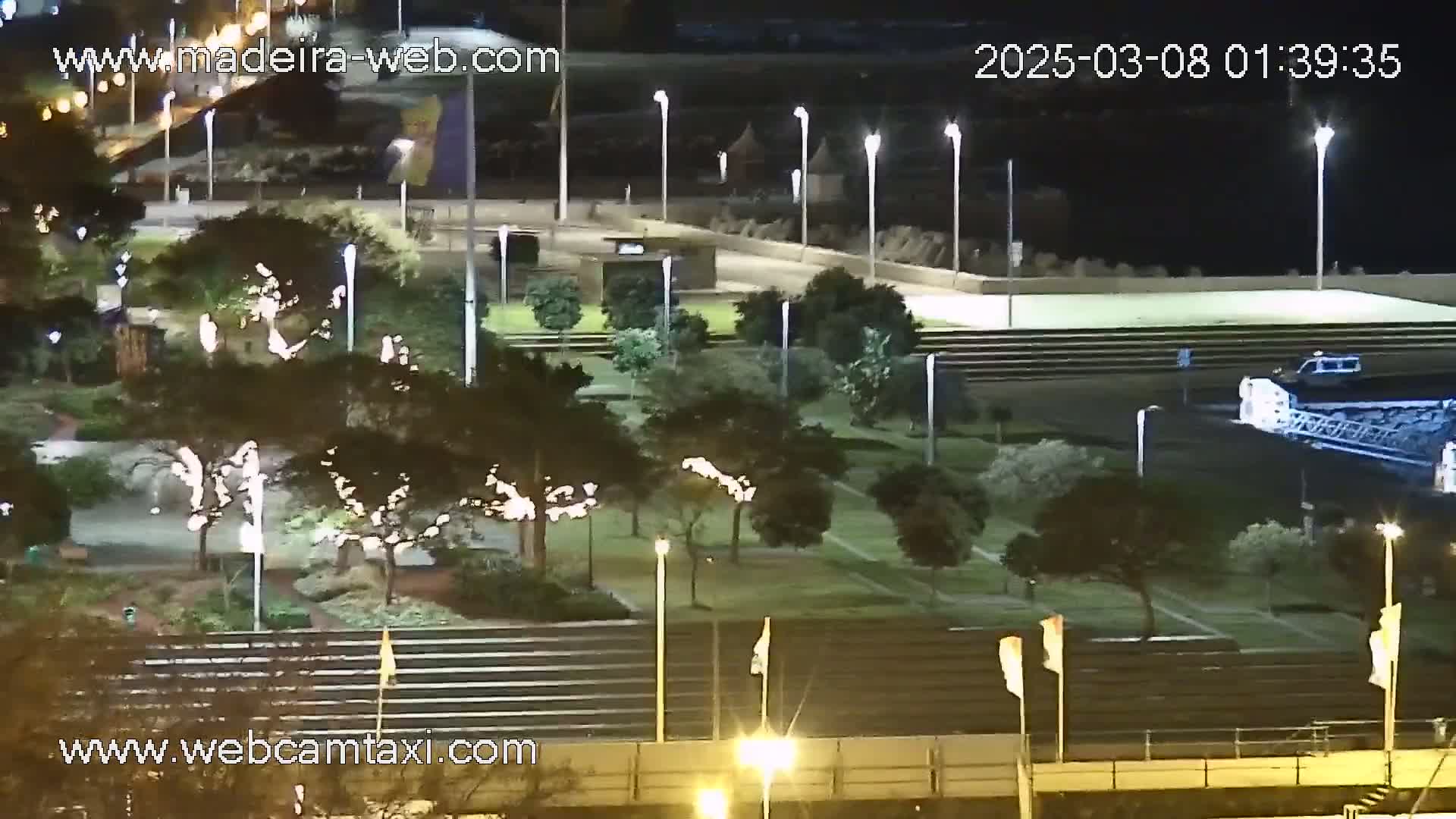 A nighttime, aerial view of a park-like area with trees and lights, next to a paved waterfront area where a vehicle is visible.