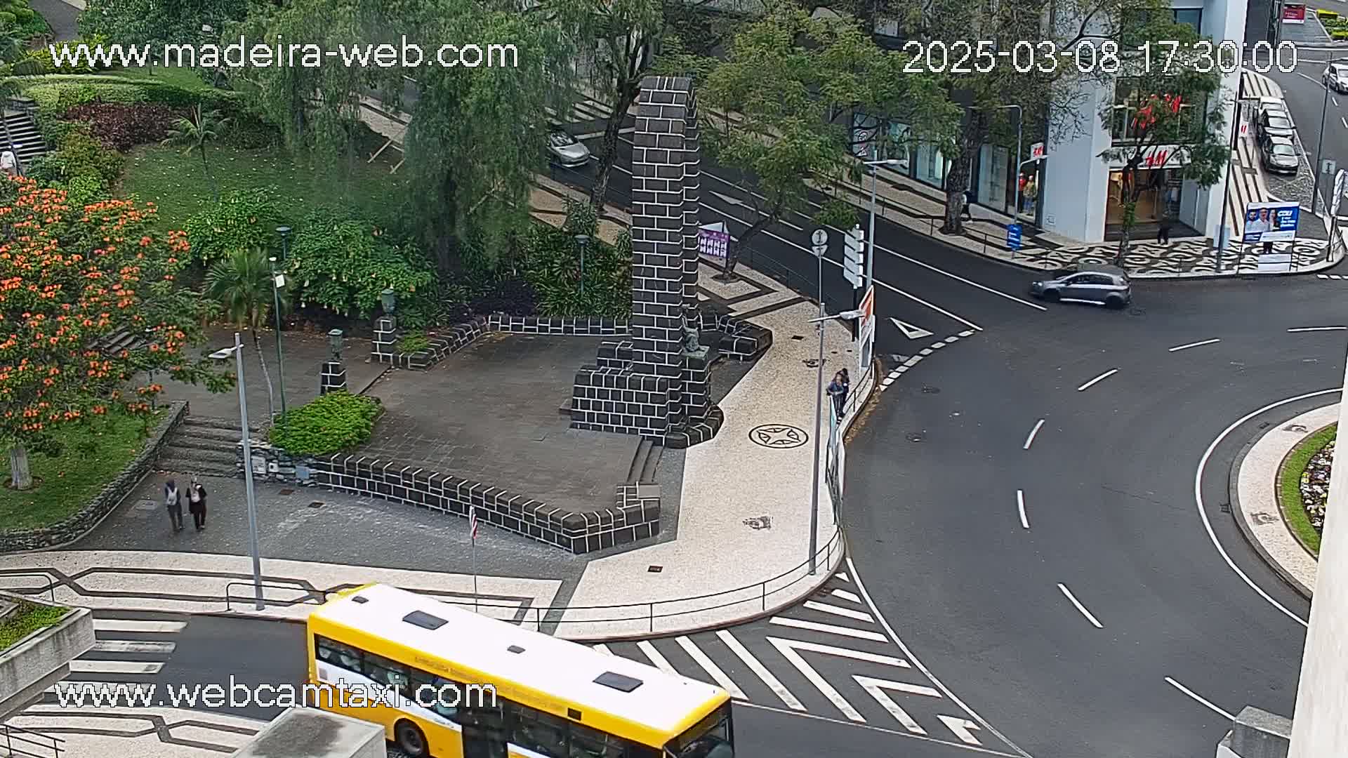 A high-angle view of a city intersection with a dark-grey paved roundabout, a small park with a dark brick monument, a yellow bus, and some pedestrians, under overcast skies.