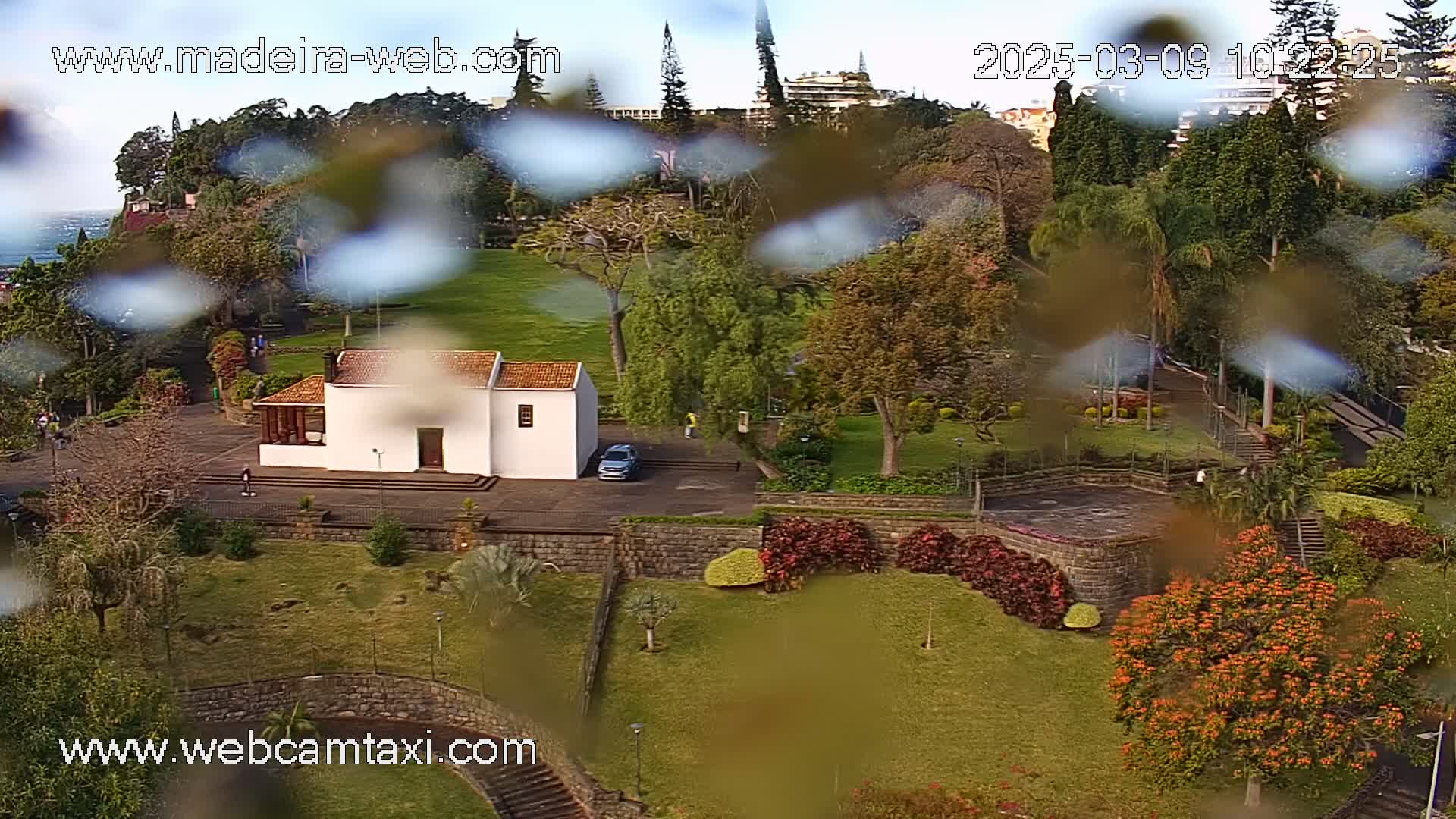 A rain-streaked view of a park with a white building, manicured lawns, and colorful landscaping, overlooking a coastline.