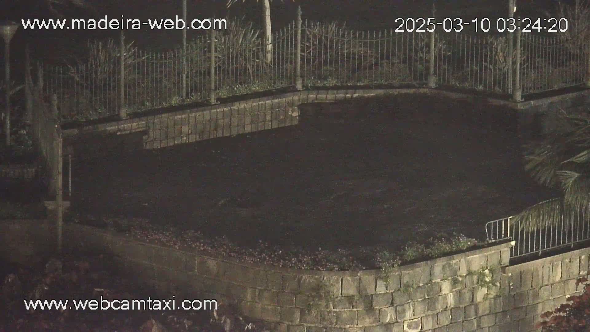 A dark, paved area is enclosed by a low stone wall and metal fence at night.