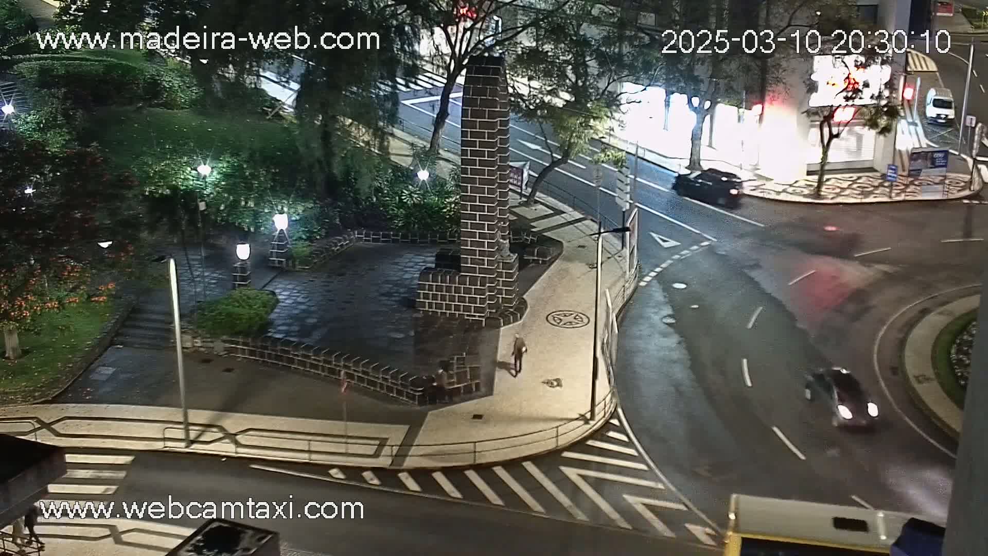 A nighttime high-angle view of a wet city intersection with a brick monument in a square, surrounded by trees and streetlights.