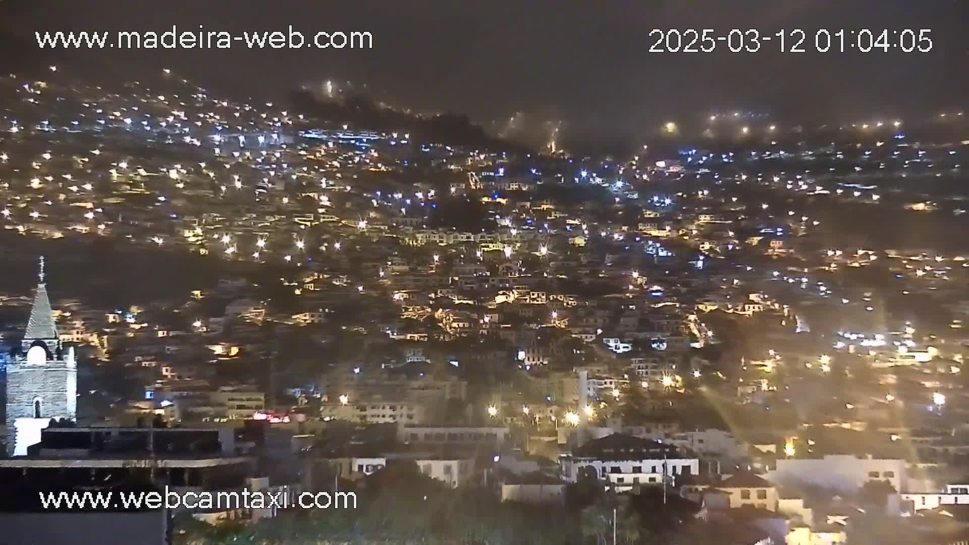A nighttime view of a densely populated hillside town, illuminated by numerous lights under hazy or foggy conditions.