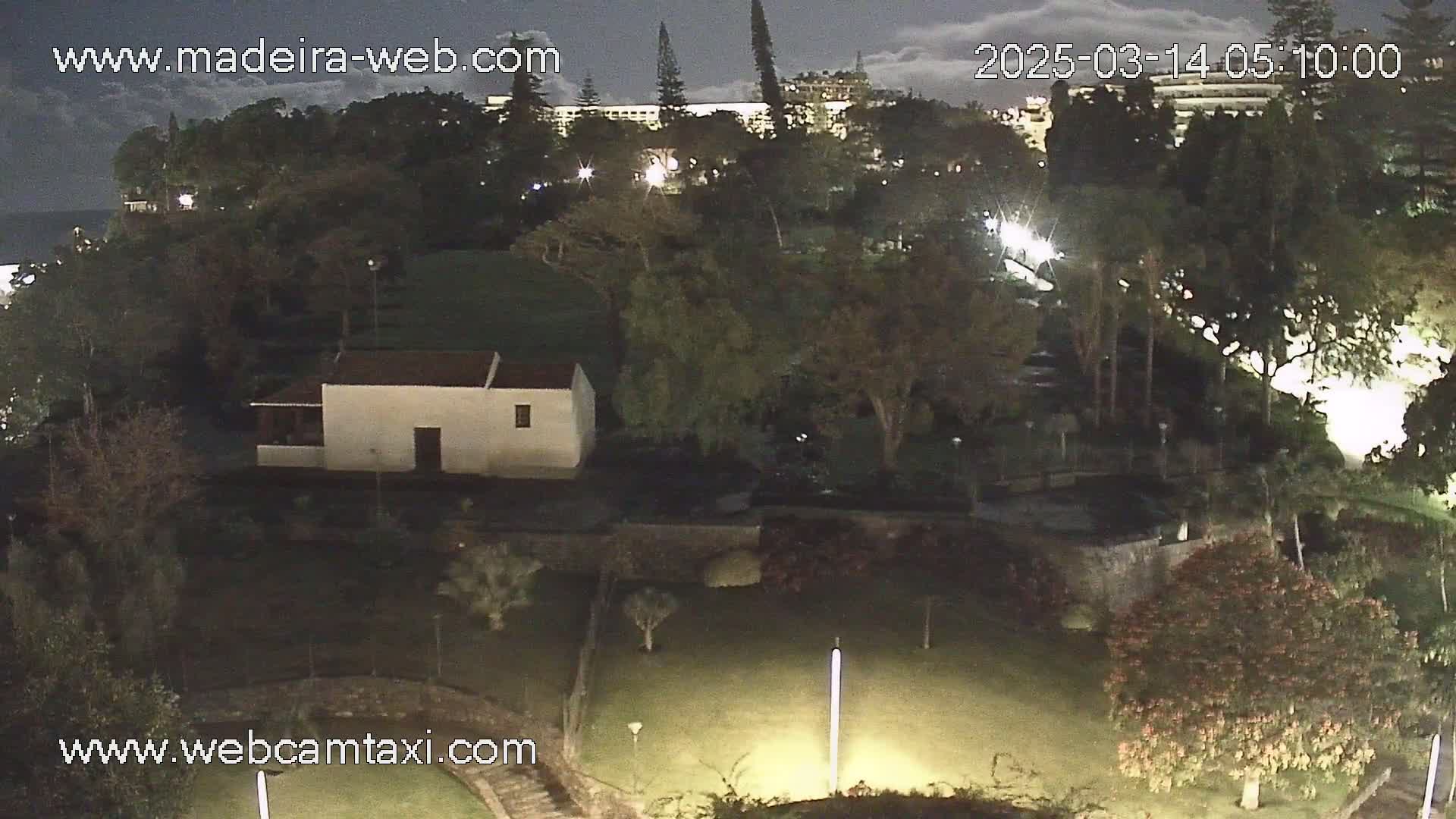 A nighttime view of a garden with a small white building, large trees, and a distant building illuminated under a partly cloudy sky.