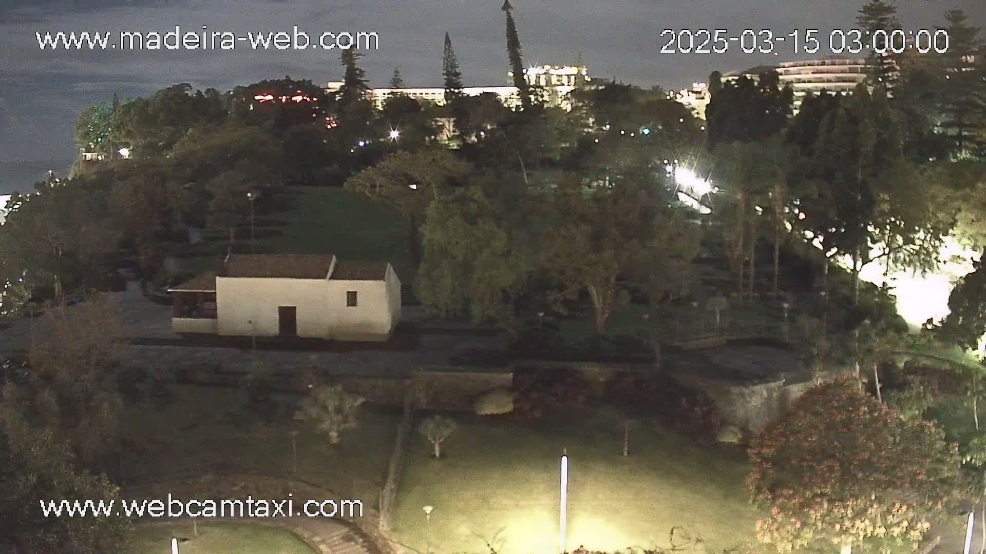 A nighttime view of a park with a small white building, lush green lawns, and trees, under a dark sky.