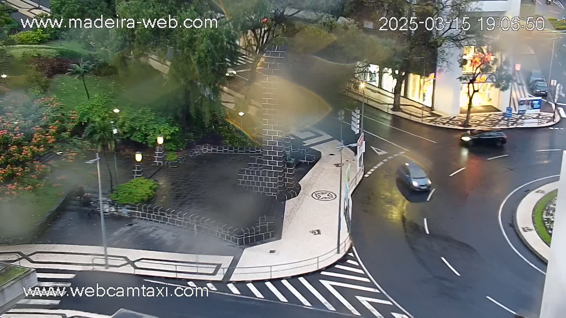 A high-angle, rainy view of a city intersection with a small plaza featuring a dark stone structure, surrounded by lush green landscaping and wet streets with several cars.