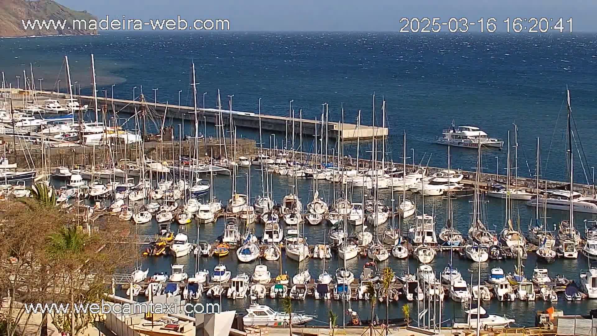 A marina filled with numerous sailboats and motorboats is seen on a sunny day with a calm sea, a larger vessel is visible further out in the water.