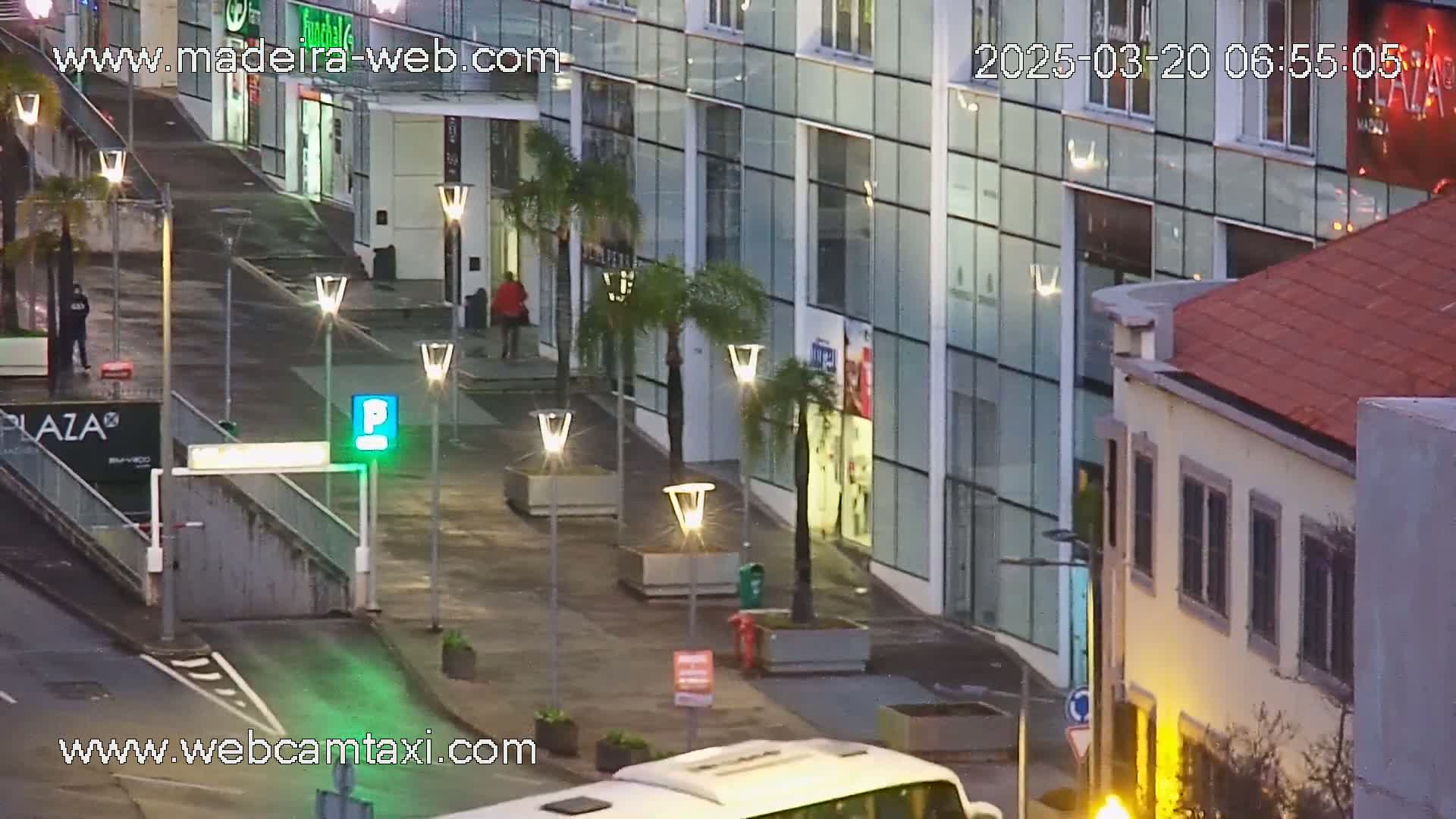A mostly empty plaza at night, under dim lighting, with a modern glass building and a smaller, older building nearby, and a bus partially visible in the foreground.