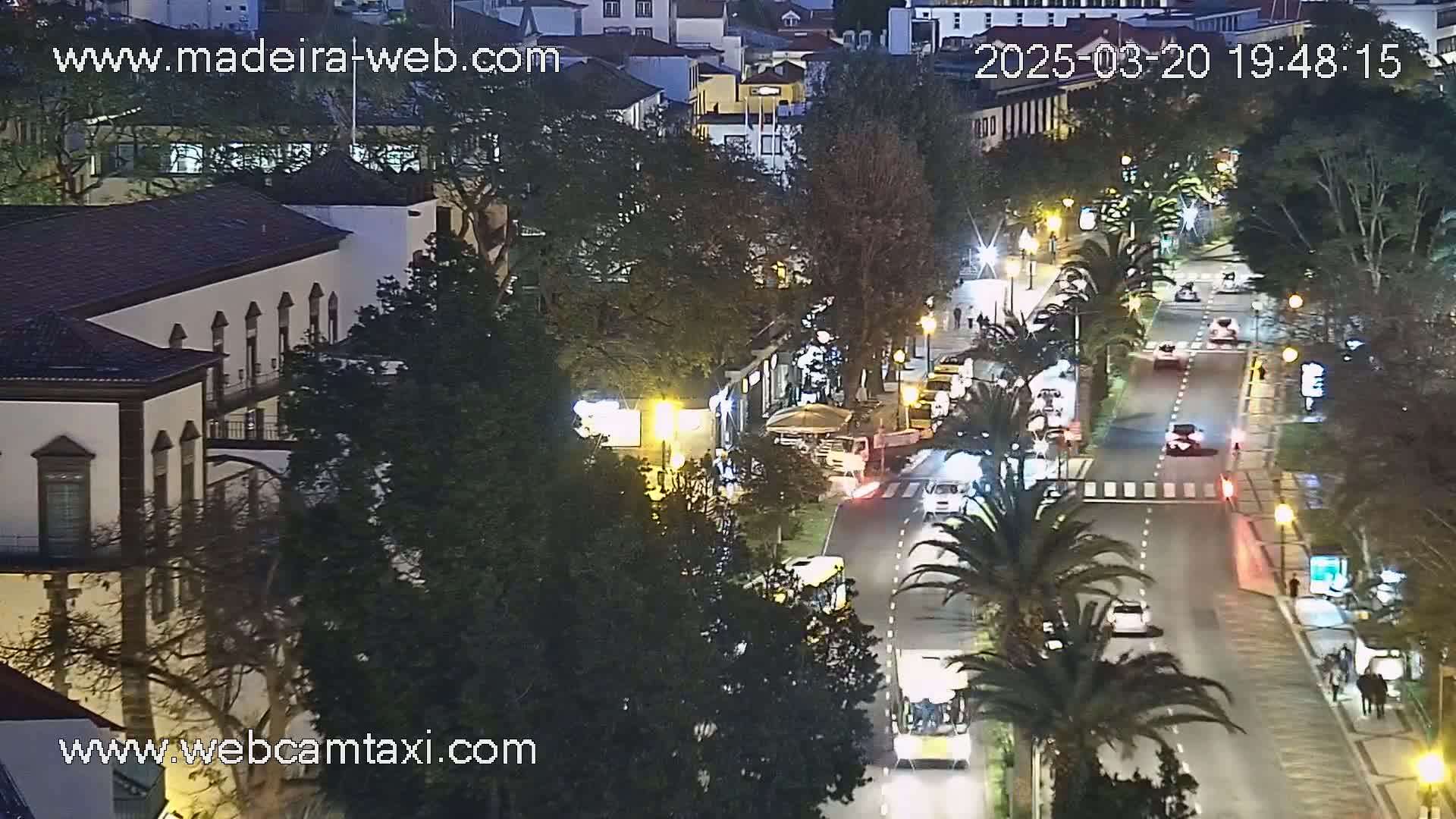 A nighttime, city street scene shows a road with cars and pedestrians under partly cloudy skies.