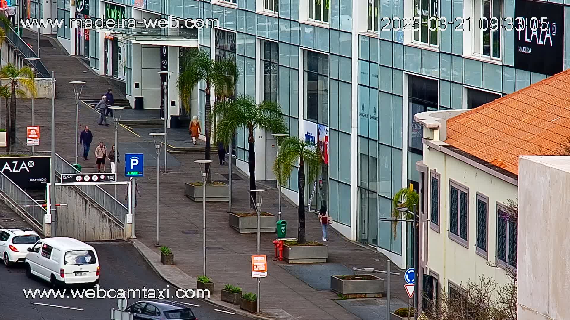 A city street scene on a sunny day shows pedestrians walking past a modern glass building and a smaller, older building with orange tile roofing.