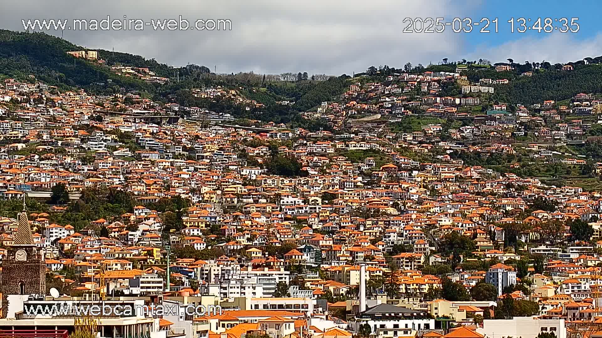 A hillside densely covered with numerous closely-packed, primarily orange-roofed buildings under a partly cloudy sky.