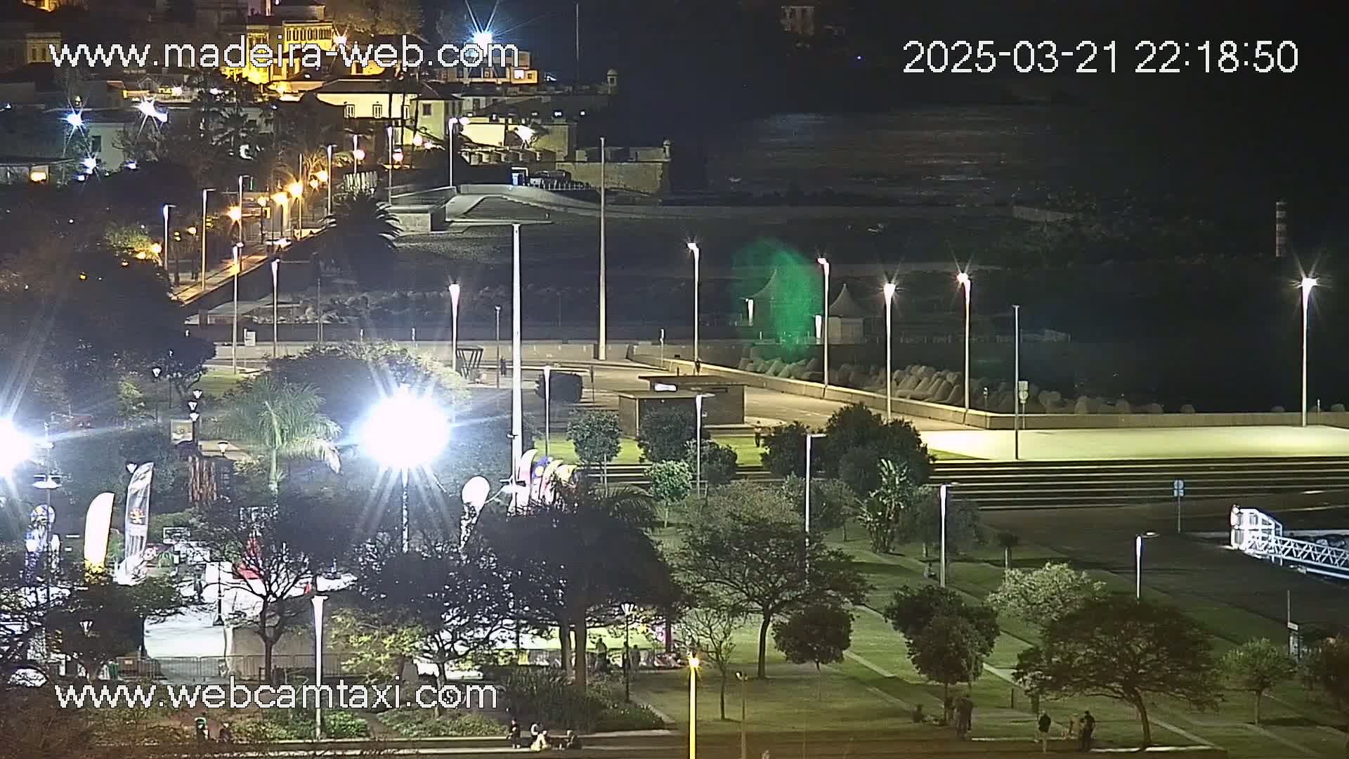 A nighttime view of a park and waterfront area, illuminated by numerous lights, with calm water visible in the background.