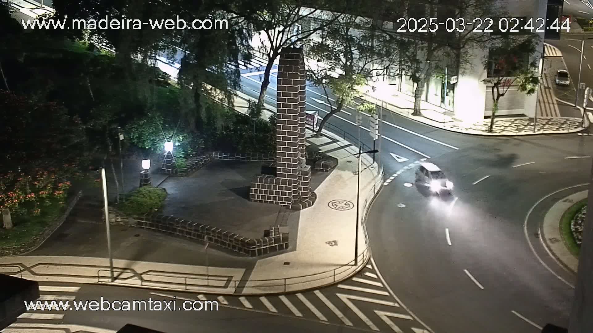 A nighttime overhead view shows a car navigating a curved road around a square plaza containing a dark brick monument, under clear skies.