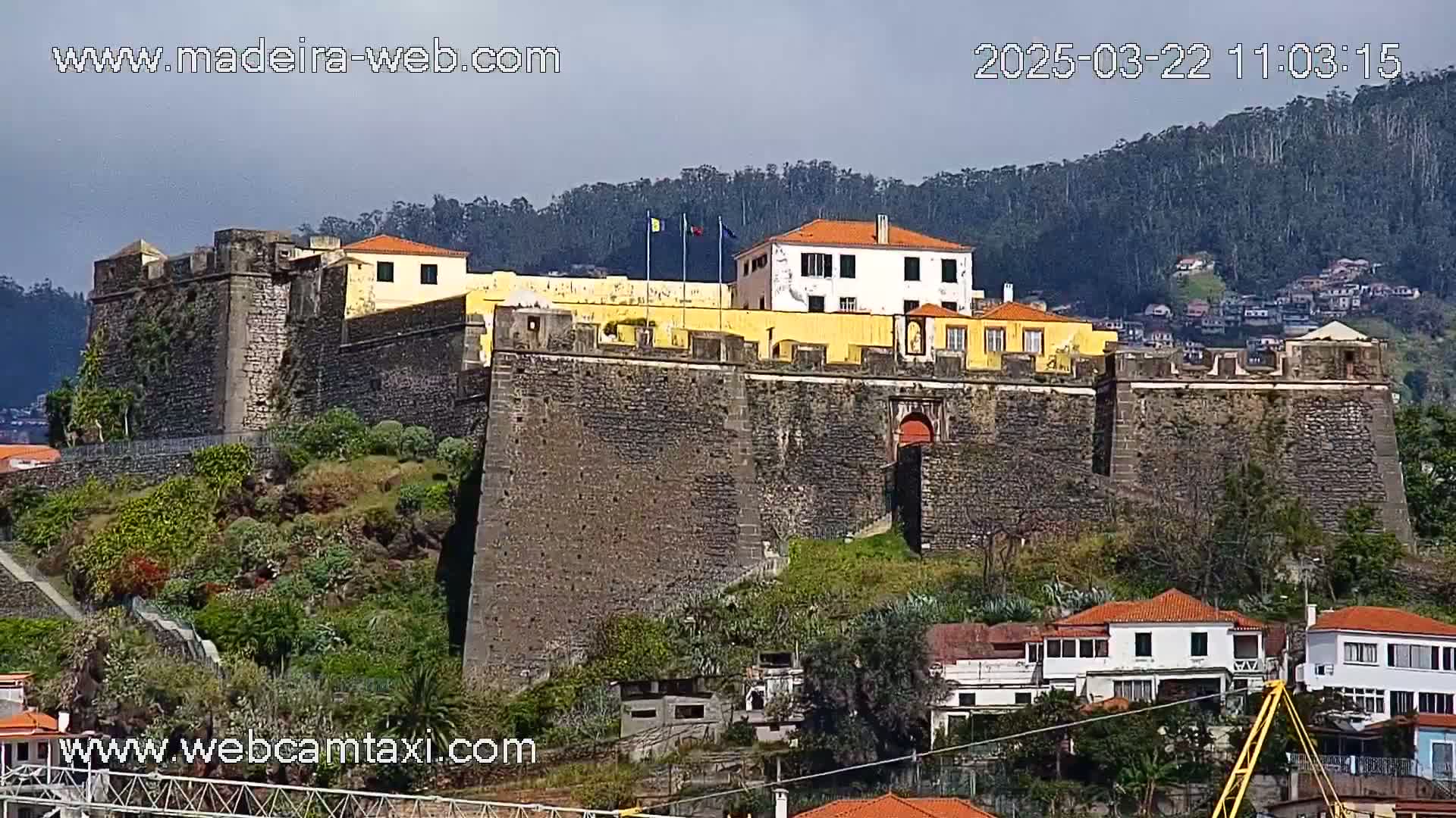 A large stone fortress with a yellow building at its center sits on a hill overlooking a town under a partly cloudy sky.