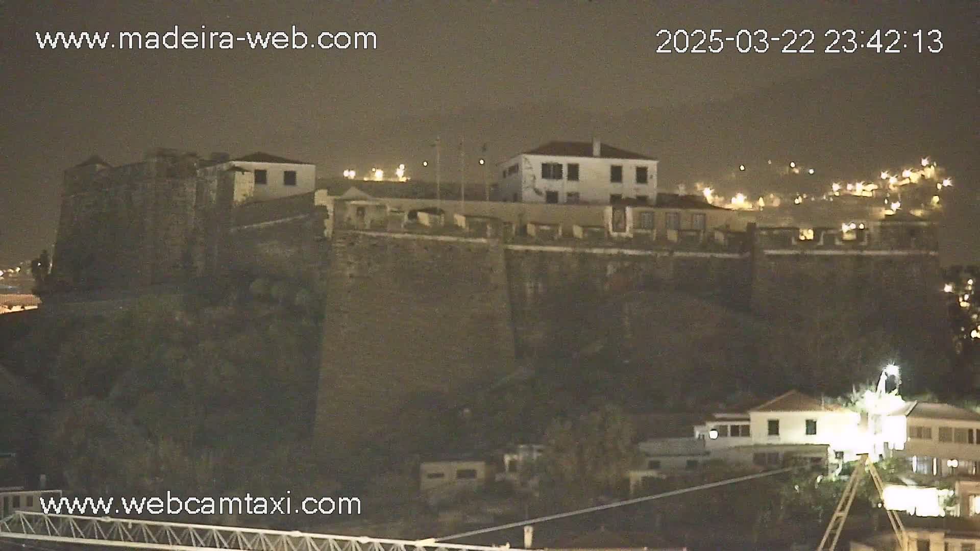 A stone fortress sits atop a hill at night, illuminated by scattered lights, under a dark, cloudy sky.