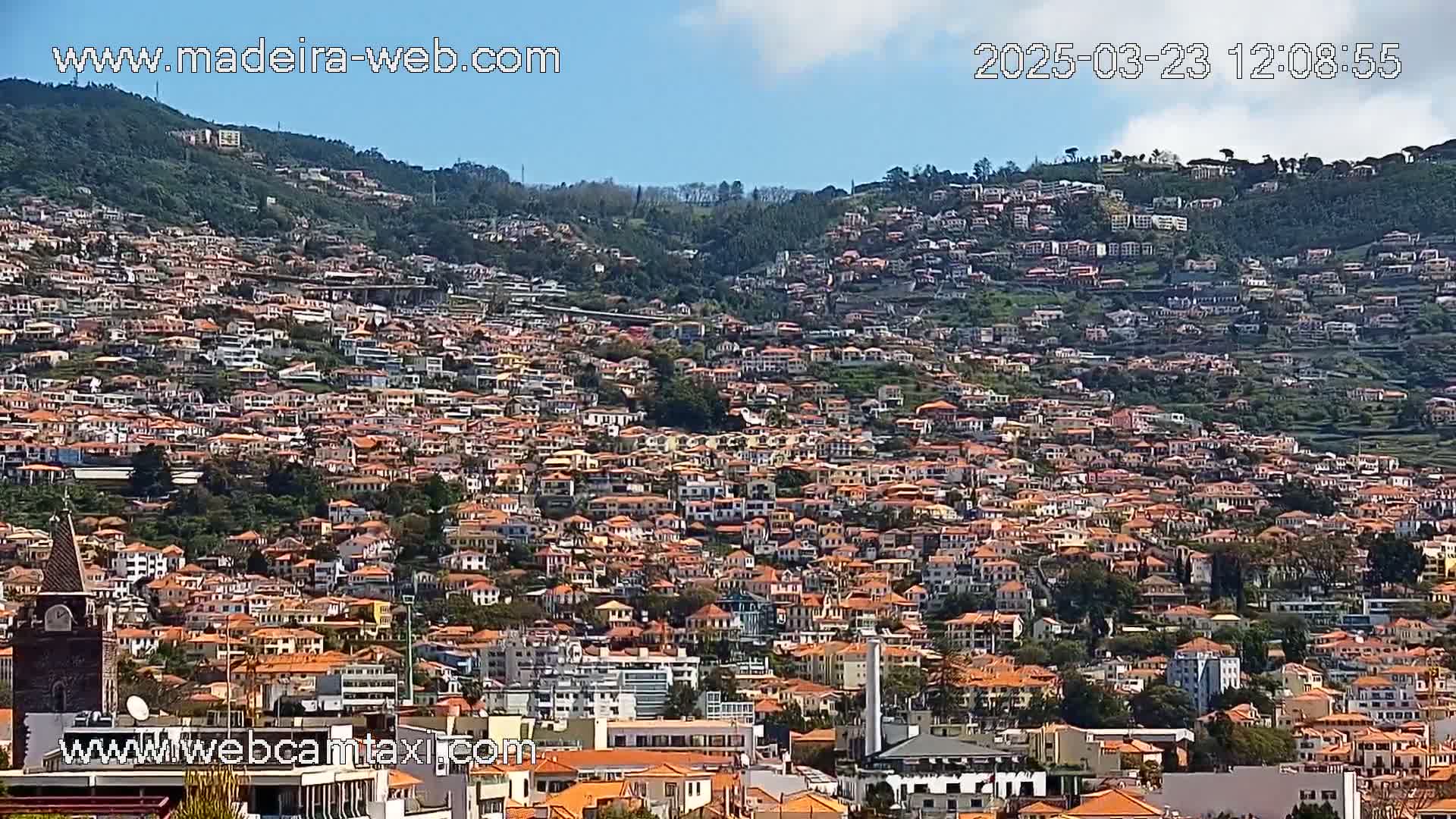 A hillside town densely packed with numerous low-rise buildings, mostly with orange roofs, under a mostly sunny sky with a few clouds.