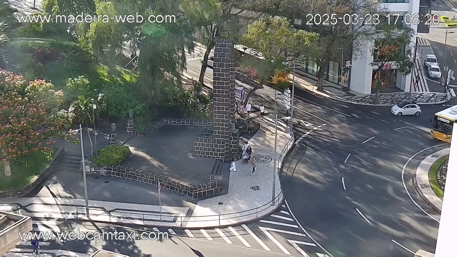 A sunny overhead view shows a city square with a dark brick monument, pedestrians, and a roadway curving around it.