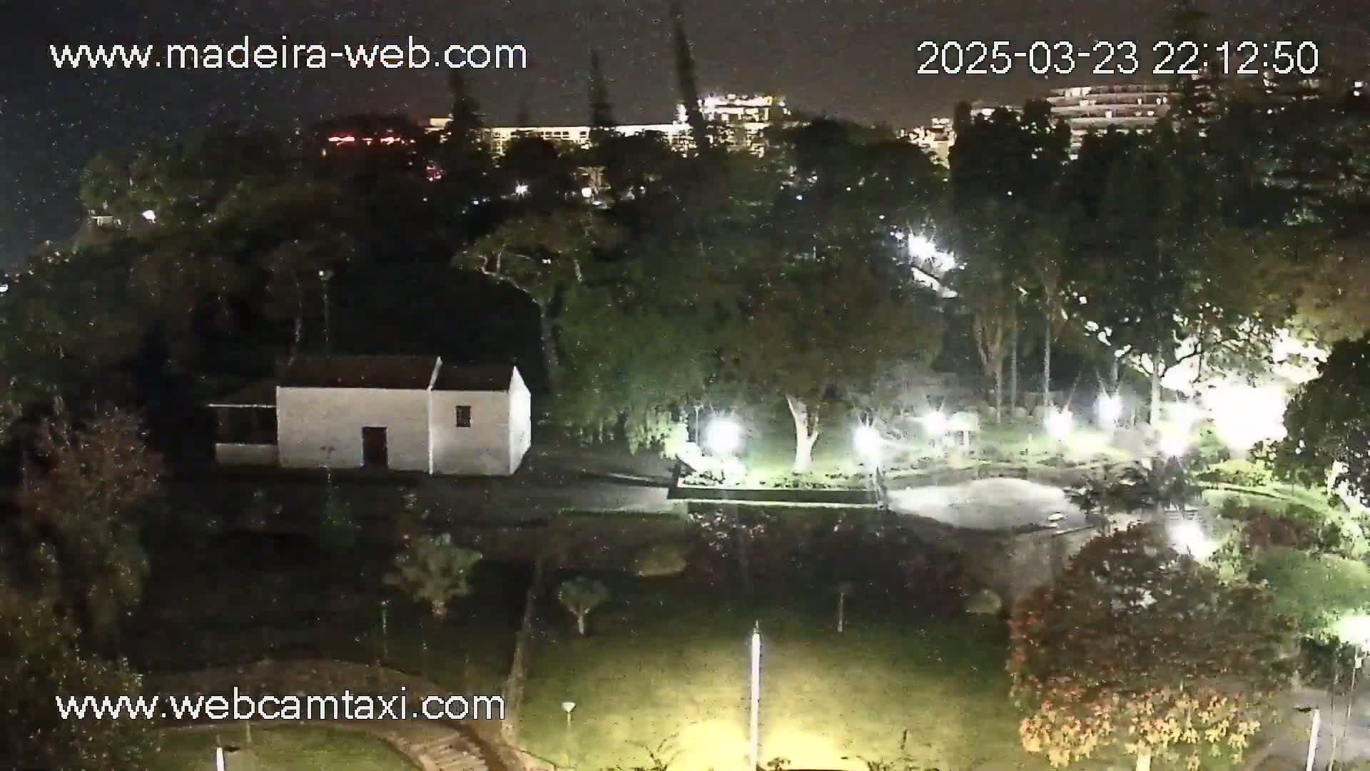 A nighttime view of a park with a small white building, illuminated by artificial lights, under slightly overcast conditions.