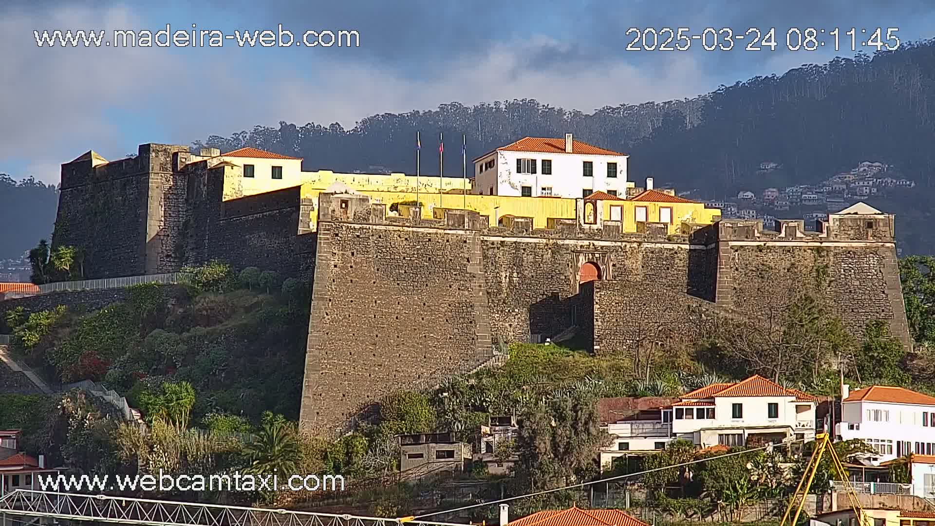 A stone fortress with a yellow building sits atop a hill overlooking a town under a partly cloudy sky.