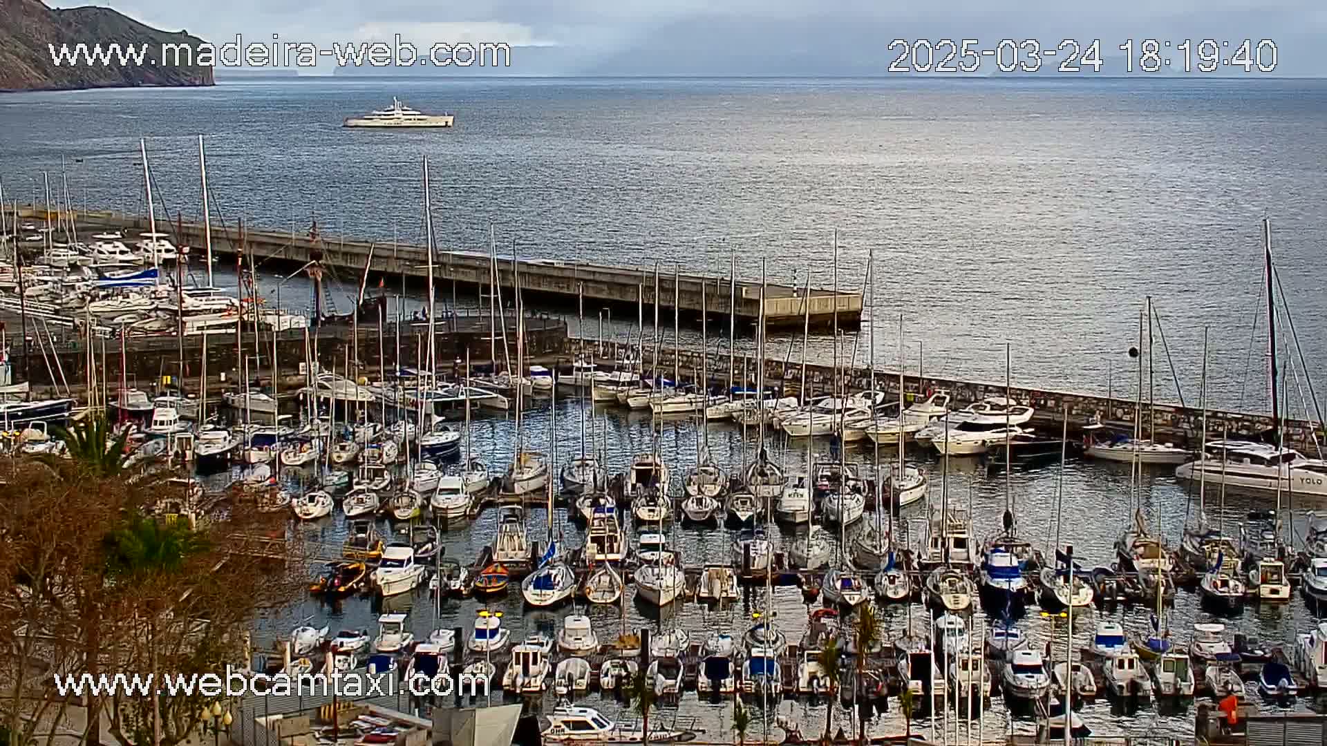 A harbor filled with numerous boats and yachts is seen on a partly cloudy day, with a larger vessel visible in the distance on open water.