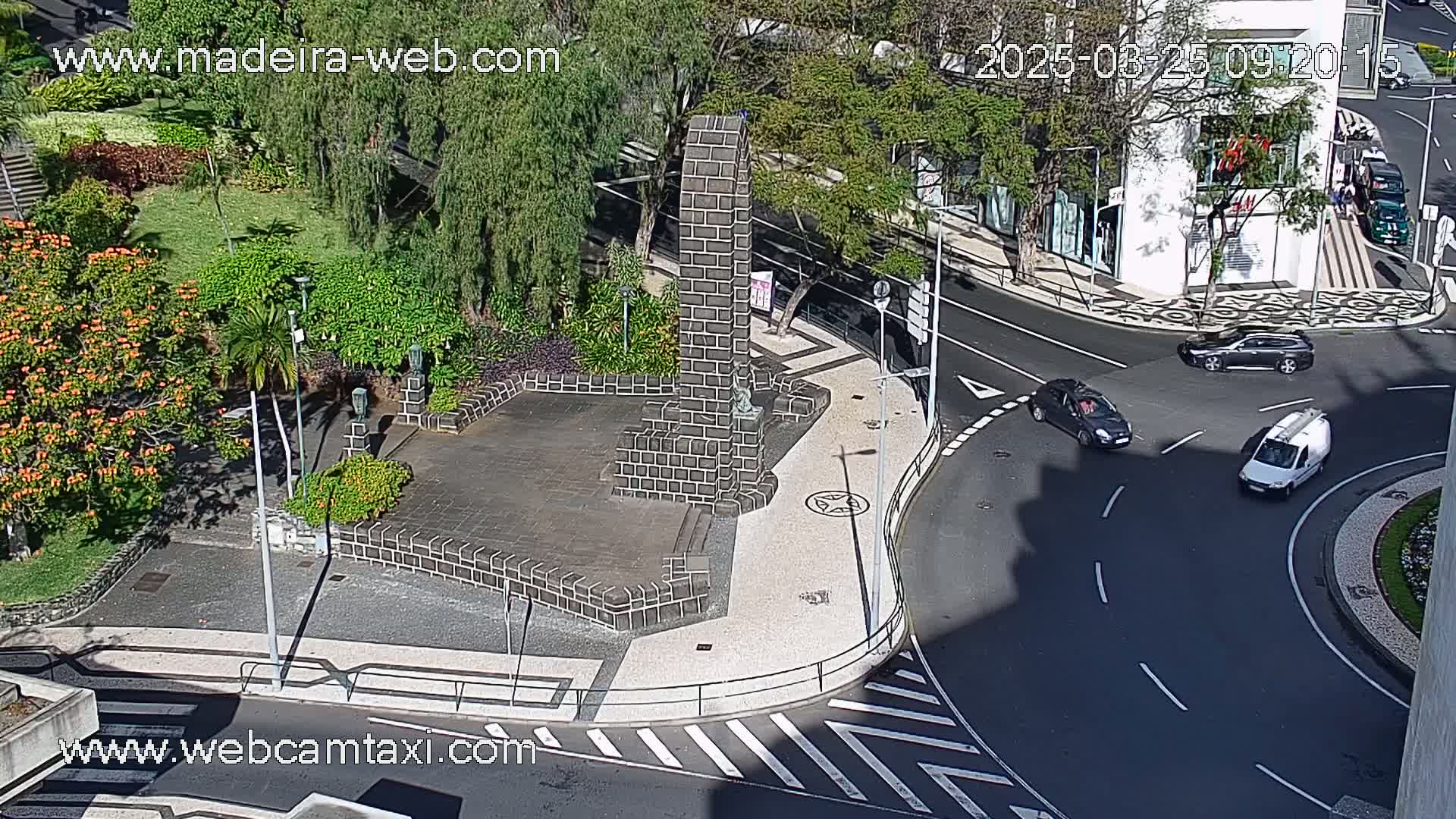 A sunny day shows a high-angle view of a paved plaza with a dark stone monument, lush green landscaping, and a roundabout with several cars.