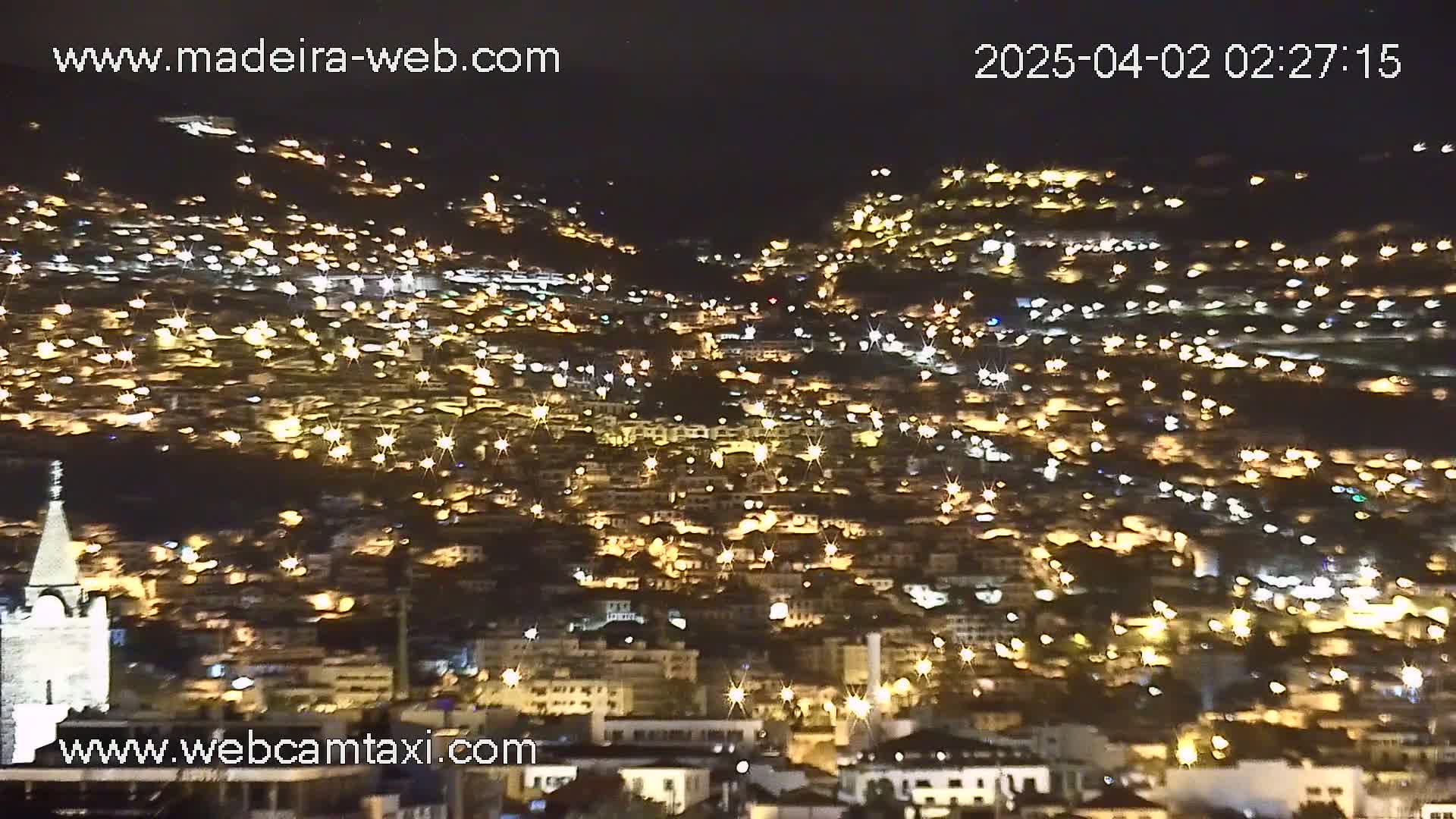 A nighttime, long shot of a city built on a hillside, illuminated by numerous lights.