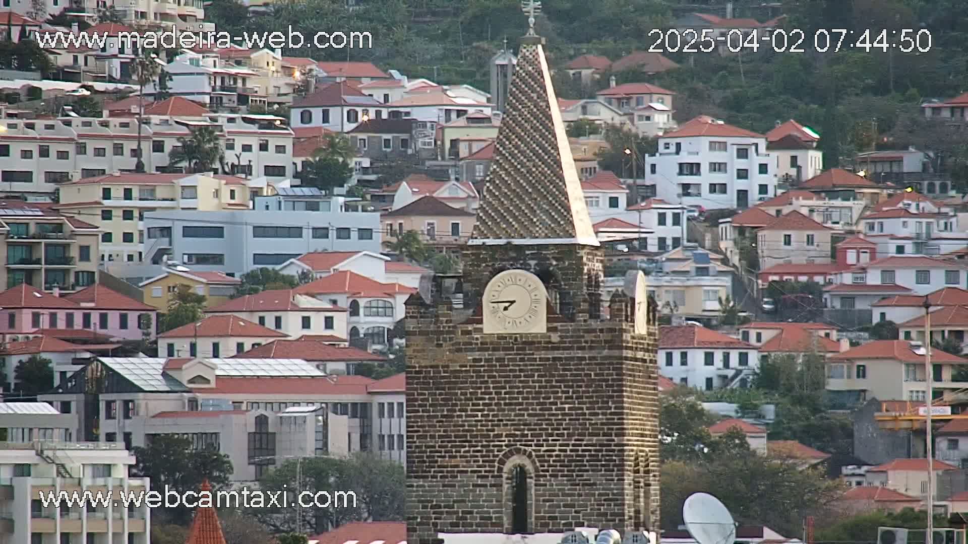 A brick clock tower stands prominently amidst a densely packed hillside of residential buildings with red-tiled roofs, under a seemingly clear sky.