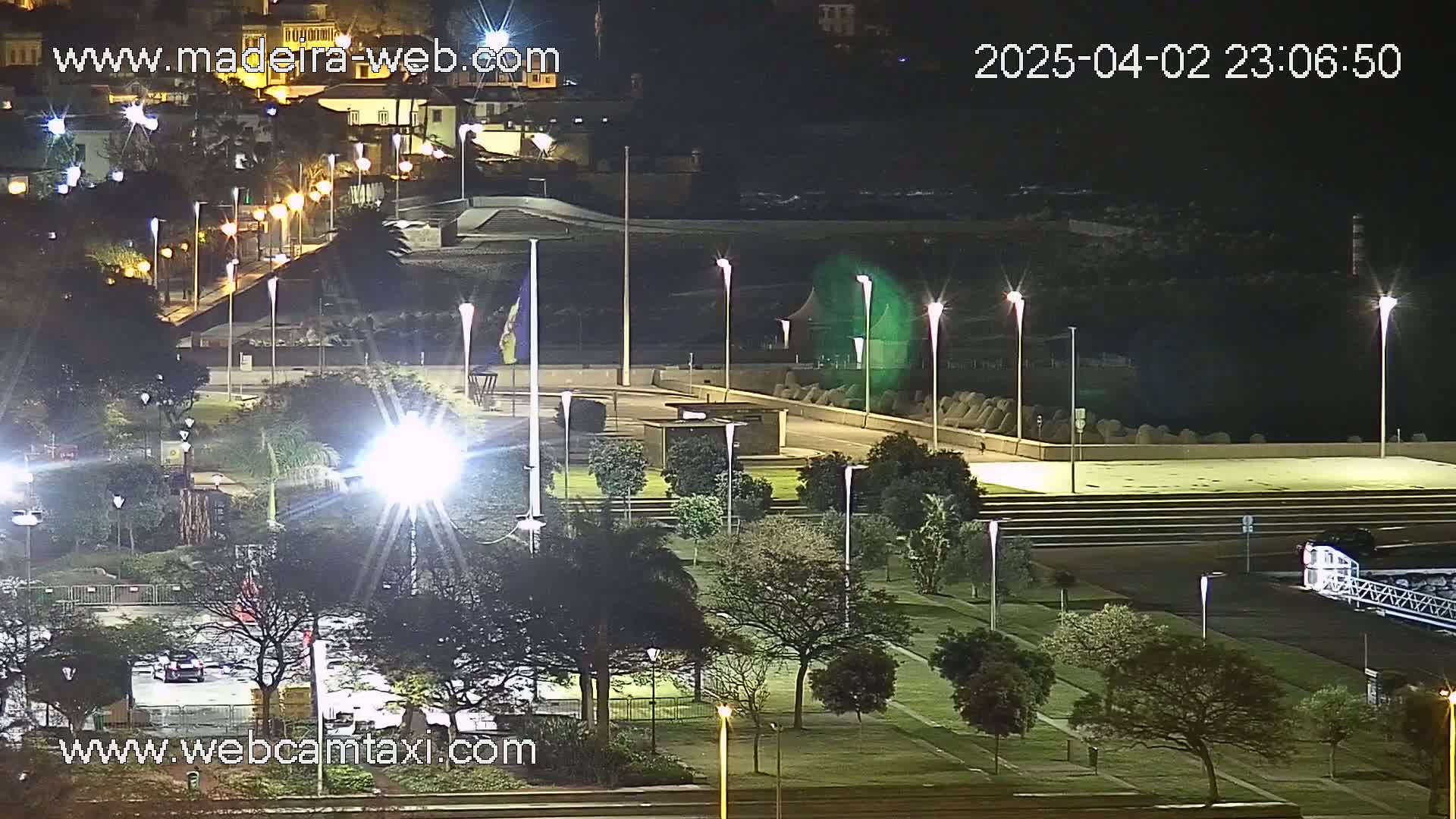 A nighttime, brightly lit park and plaza overlook a dark body of water, with a town visible in the distance.