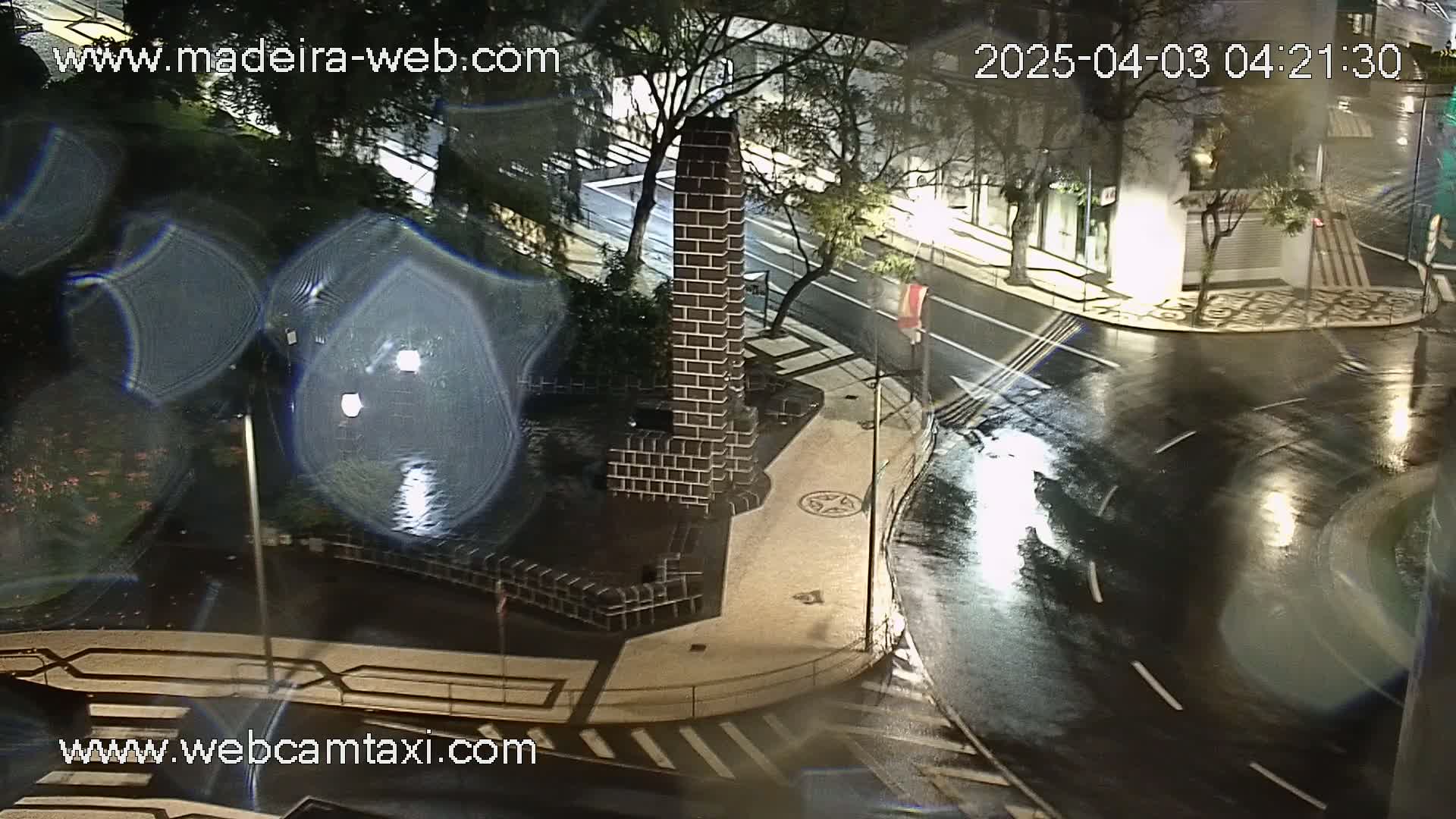 A brick monument is situated at a rain-slicked intersection at night, with streetlights reflecting on the wet pavement and cars moving in the distance.
