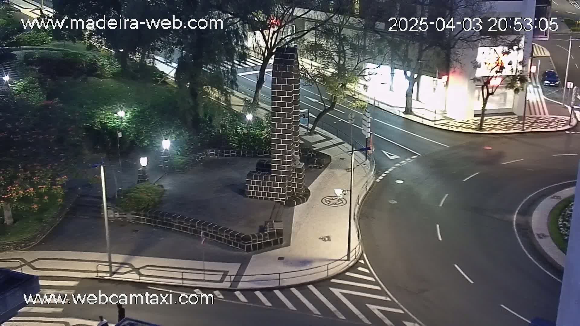 A nighttime aerial view shows a dark brick monument in a small plaza surrounded by trees and streetlights, next to a curved roadway.