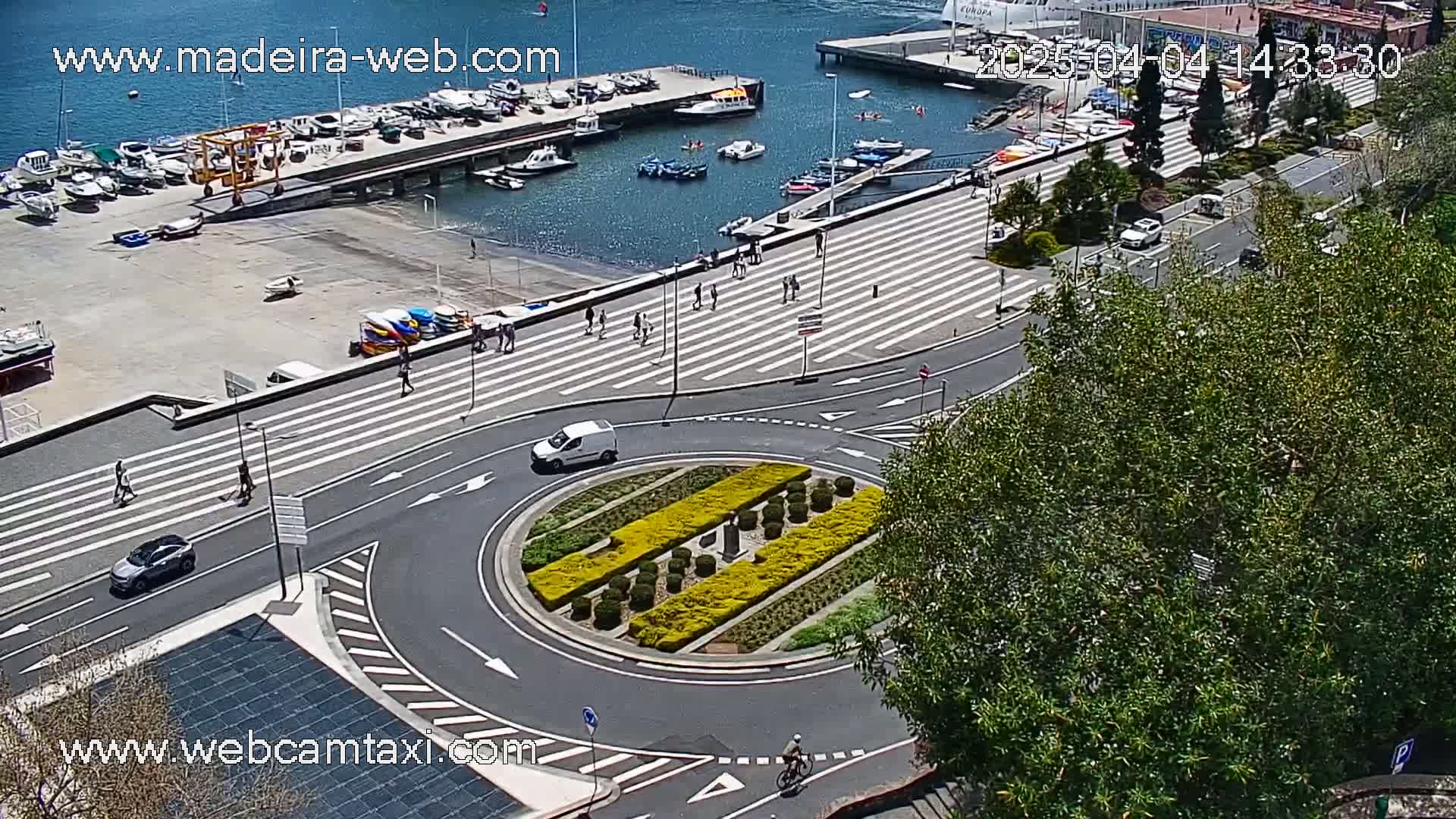 A high-angle view shows a harbor with docked boats, a roadway with a roundabout, pedestrians walking on a crosswalk, and several vehicles driving, all under sunny conditions.