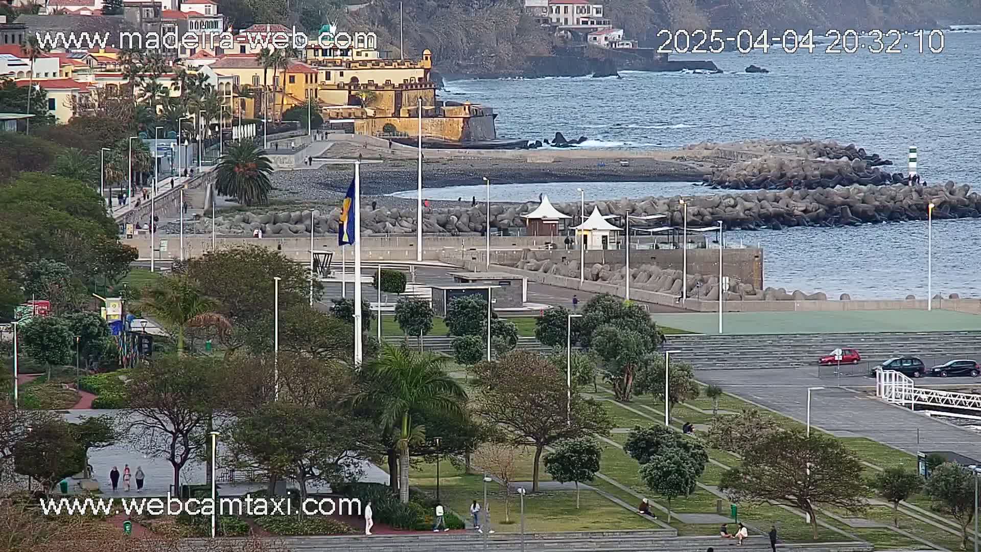 A coastal park with a paved area, green lawns, trees, and a few people walking, overlooks a calm ocean with a breakwater and a distant town under a mostly clear sky.