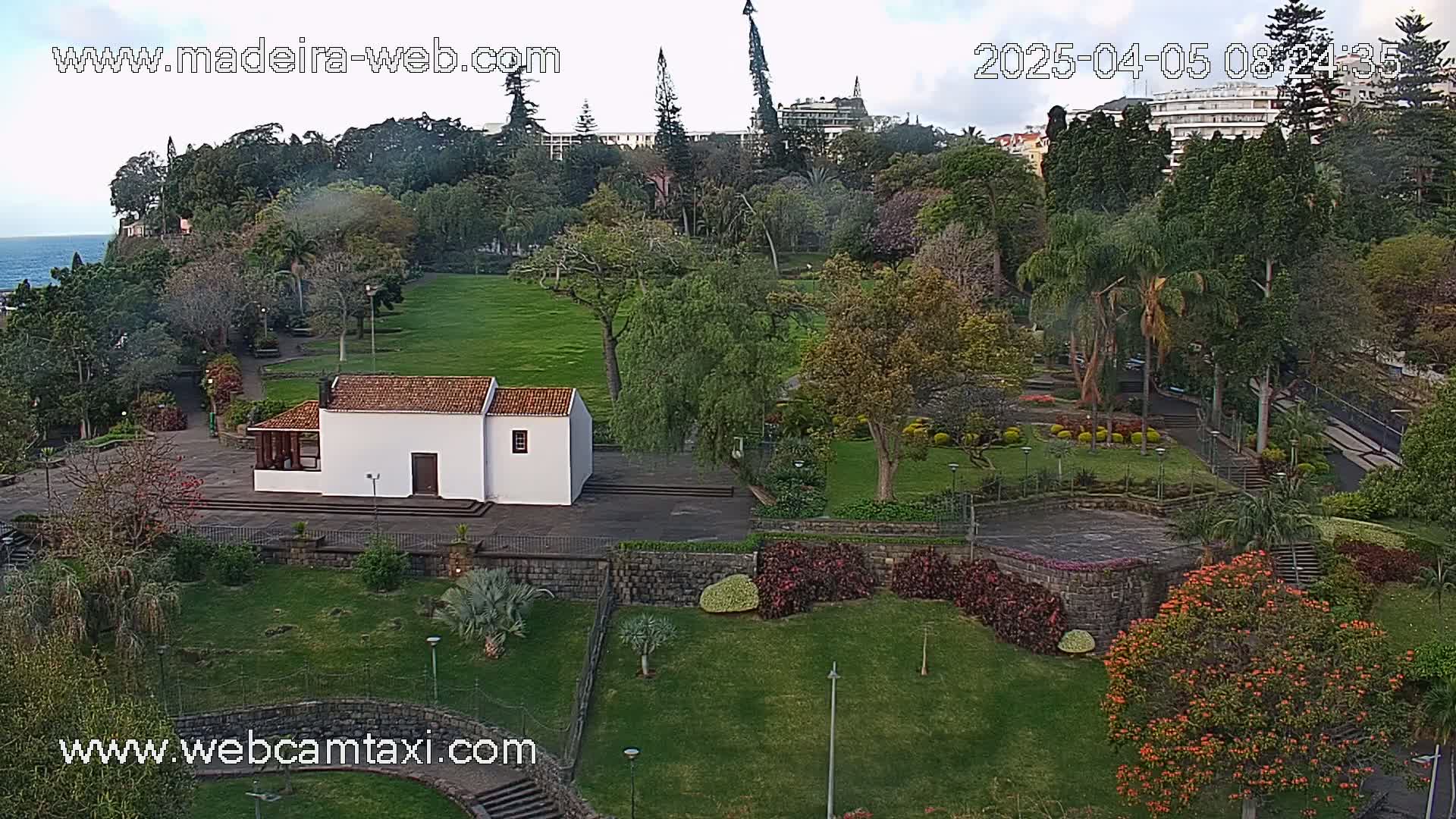 A verdant park with a small white building, various trees and shrubs, and a stone wall overlooks the ocean under a partly cloudy sky.