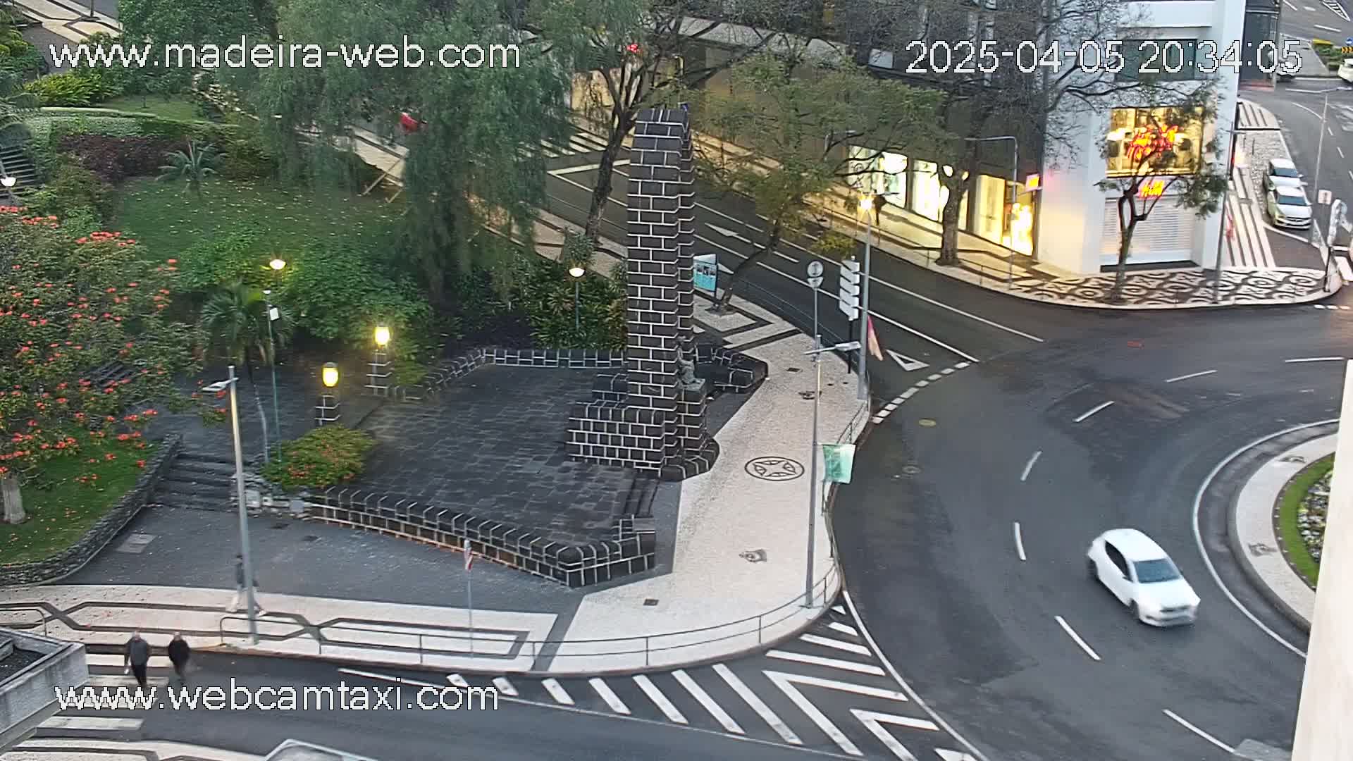 A high-angle view shows a paved plaza with a dark brick monument and surrounding walkways, next to a street intersection with a car driving around a curve at night.