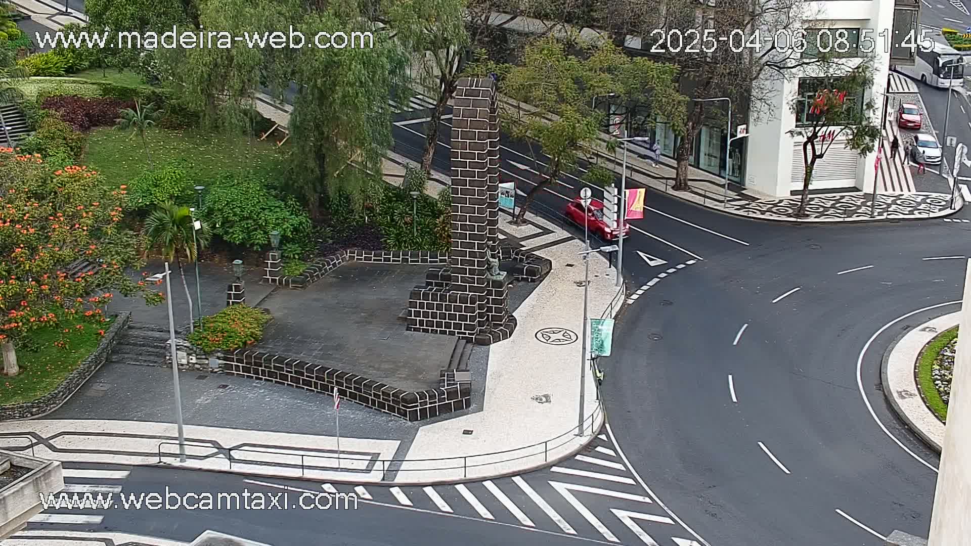 A brick monument stands in a paved plaza next to a curved roadway in a city, with lush green landscaping visible in the background.