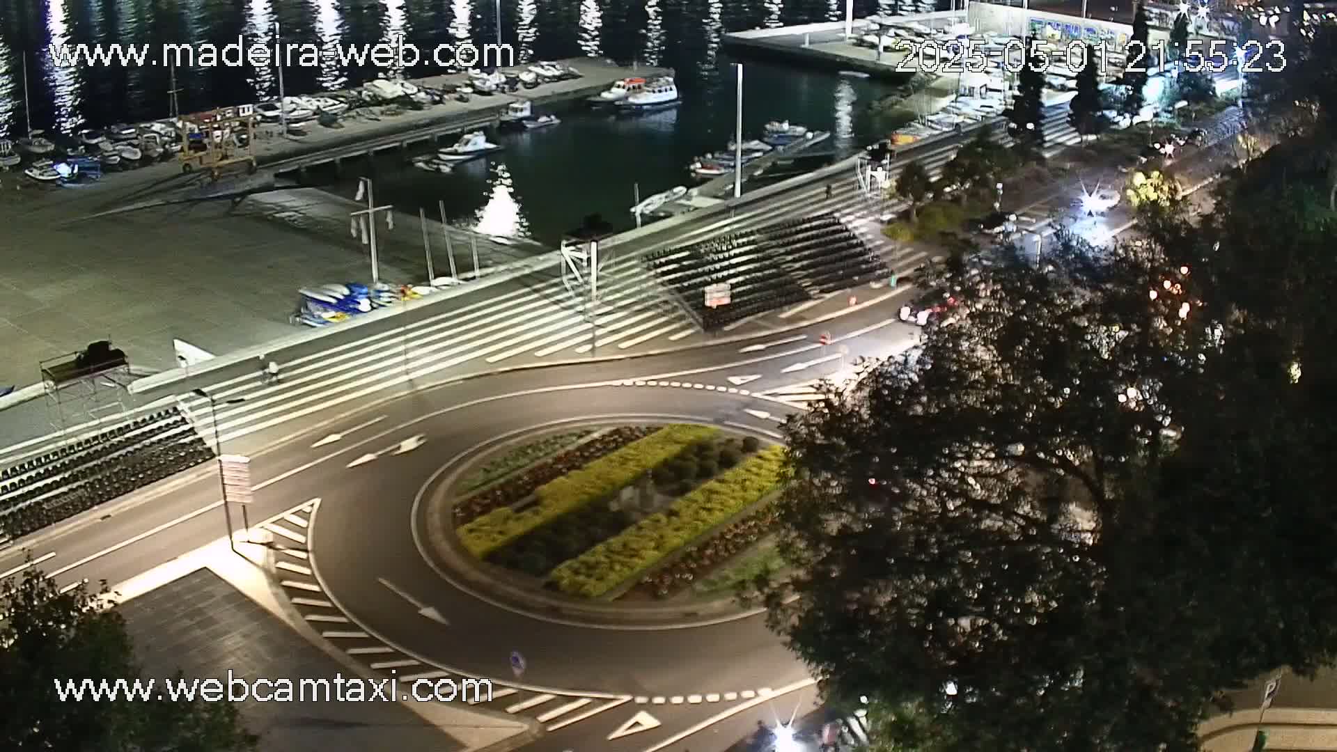 A nighttime aerial view shows a harbor with docked boats, a roundabout with landscaping, and a roadway with cars, all under a dark sky.