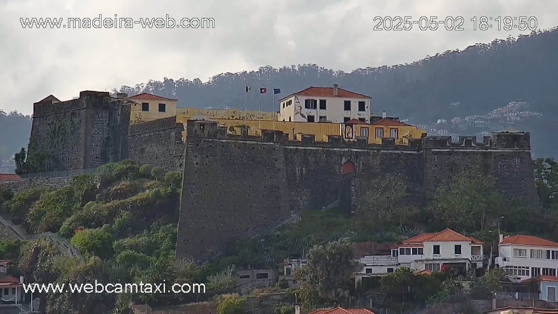 A large stone fortress sits atop a hill overlooking a town under an overcast sky.