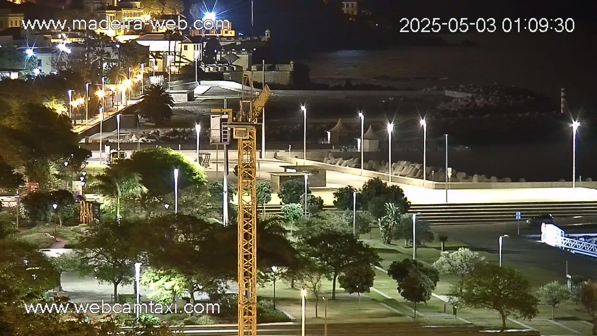 A nighttime view of a park-like area next to a body of water, illuminated by numerous lights, with a construction crane visible and trees surrounding a paved area.