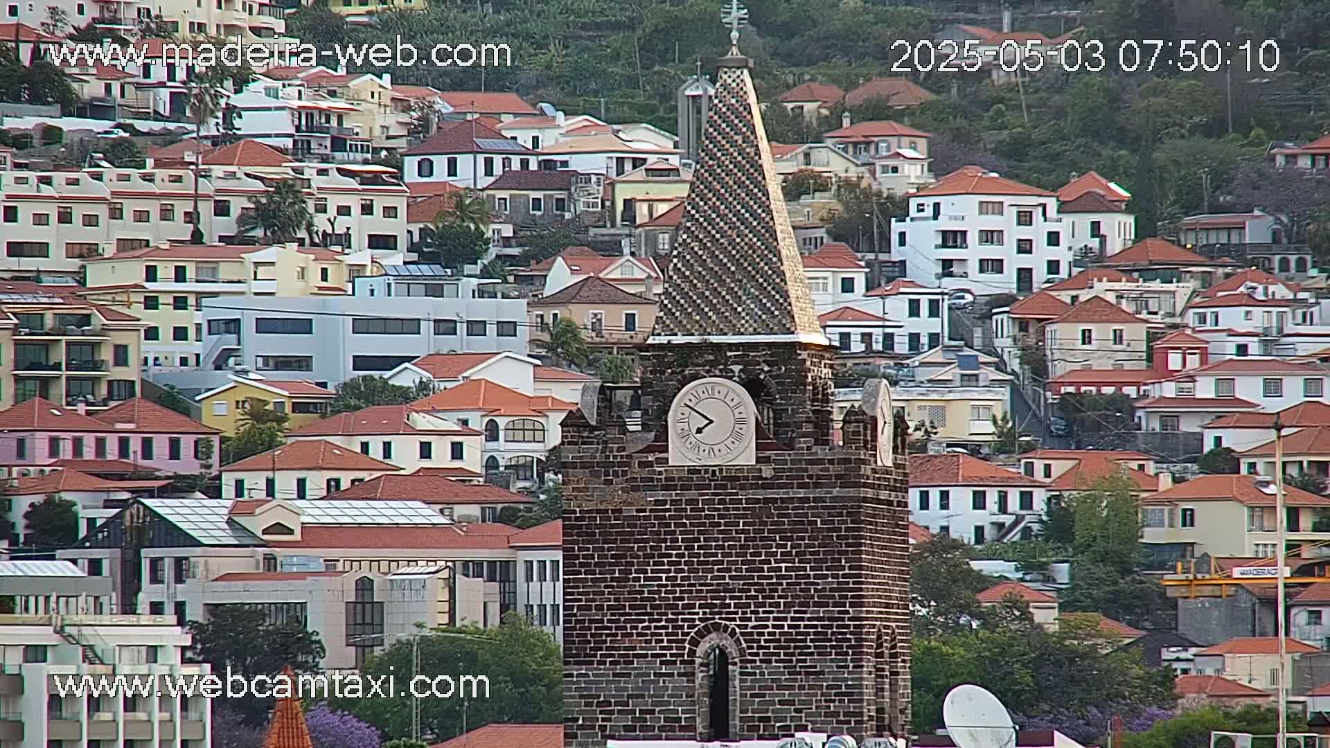 A brick clock tower with a tiled spire stands prominently among a dense cluster of red-roofed buildings on a hillside, under an overcast sky.
