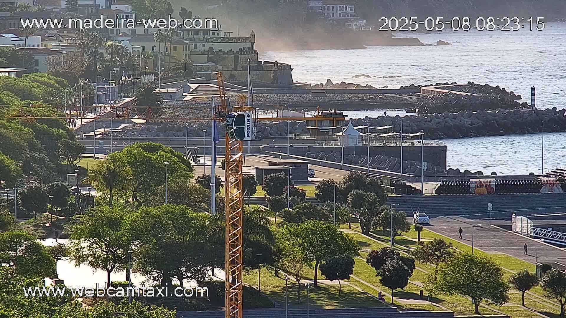 A coastal scene on a sunny day shows a construction crane, a park with lush green trees, a paved walkway along the ocean, and a rocky breakwater protecting a calm sea.