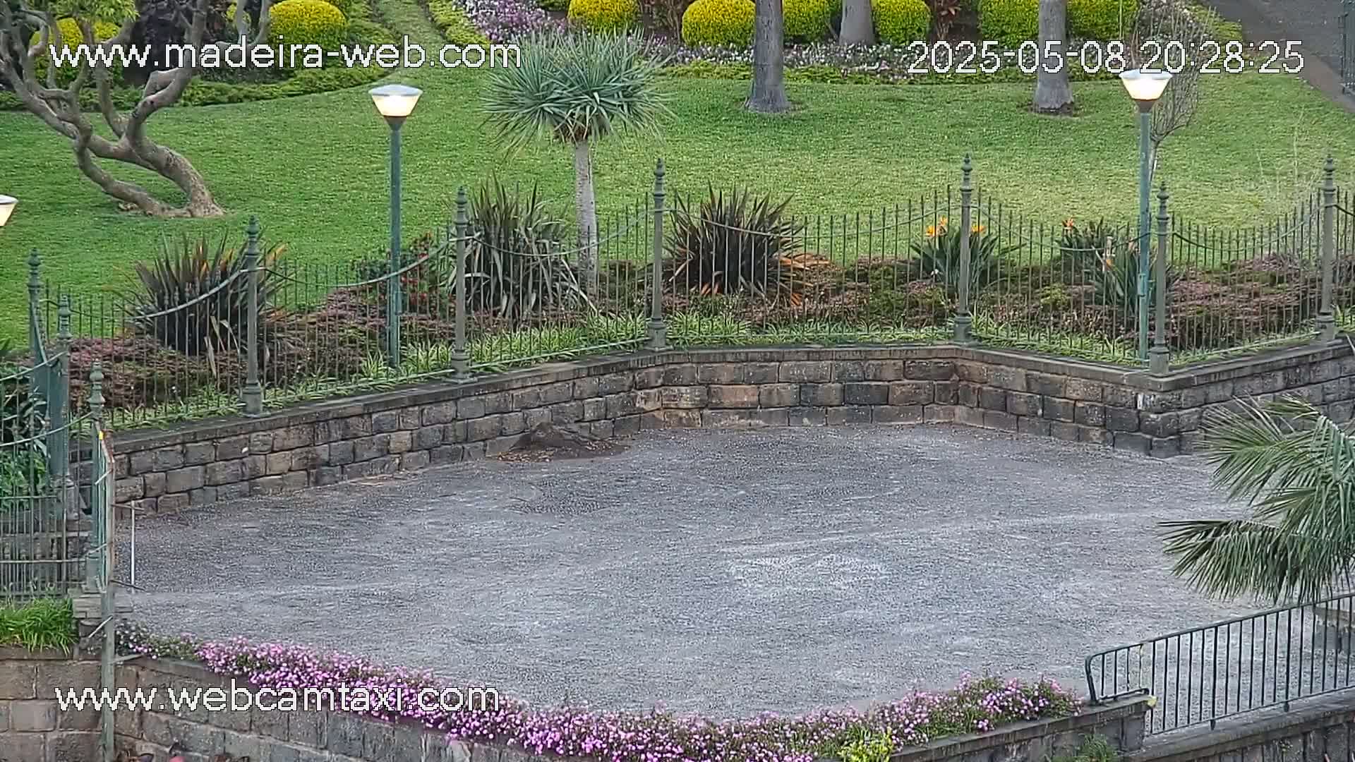 A wet, paved area is bordered by a low stone wall and iron fencing, encompassing lush green lawns, flowering plants, and palm trees under an overcast sky.