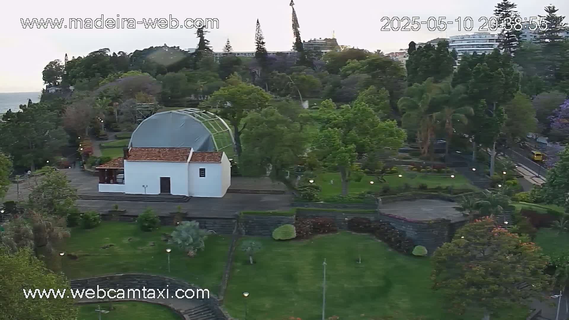 An aerial view shows a white building with a reddish-brown roof in a lush green park, overlooking a calm ocean under an overcast sky.