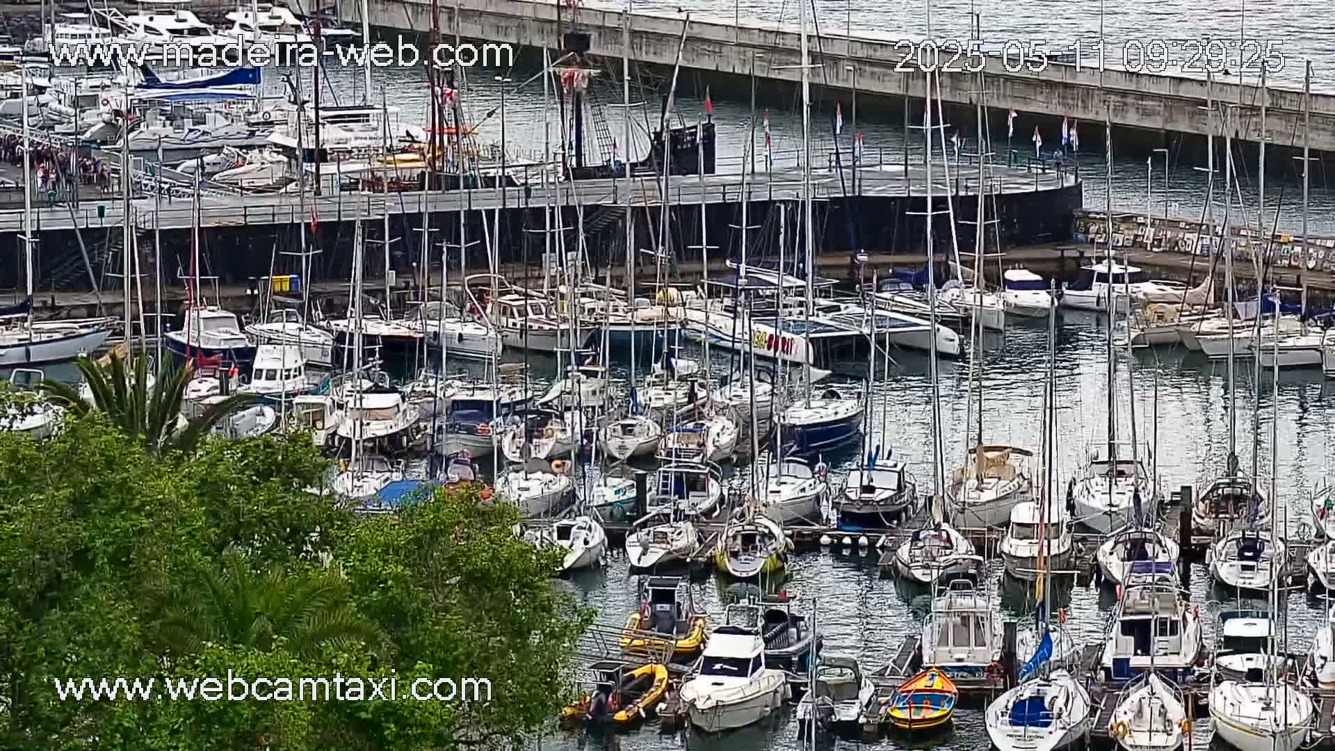 A marina filled with numerous sailboats and motorboats is viewed from above on a partly sunny day.