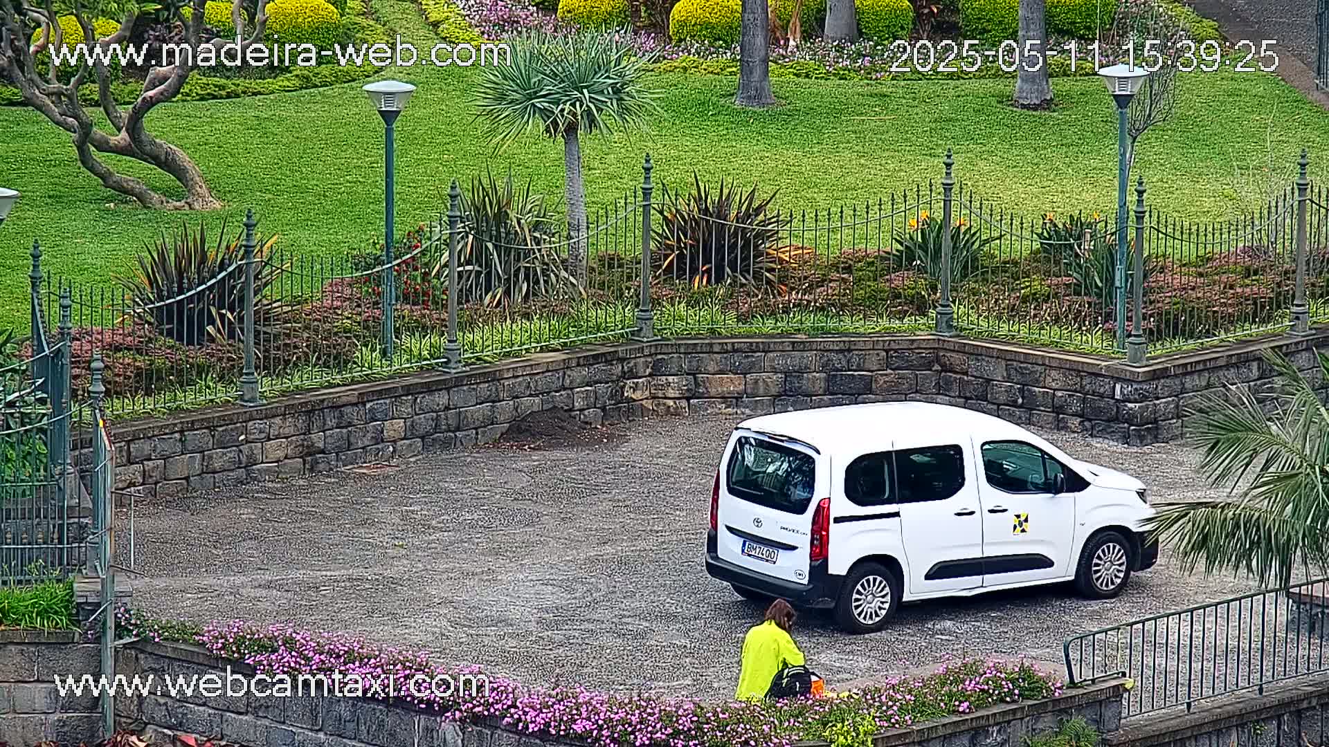 A white van is parked in a gravel area next to a stone wall and lush green lawn, bordered by wrought-iron fencing and flowering plants, under an overcast sky.